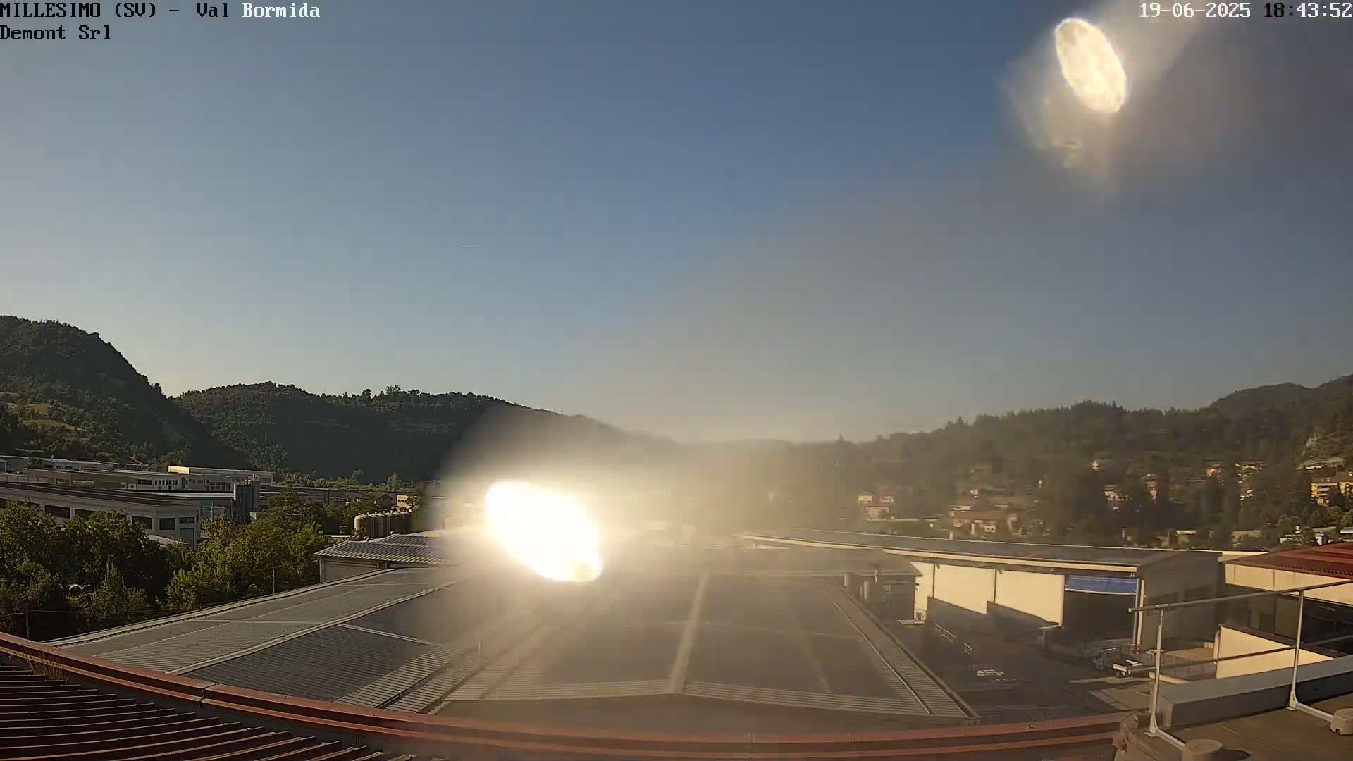 A hazy sun shines brightly over a landscape of green hills and industrial buildings, with a clear blue sky above.
