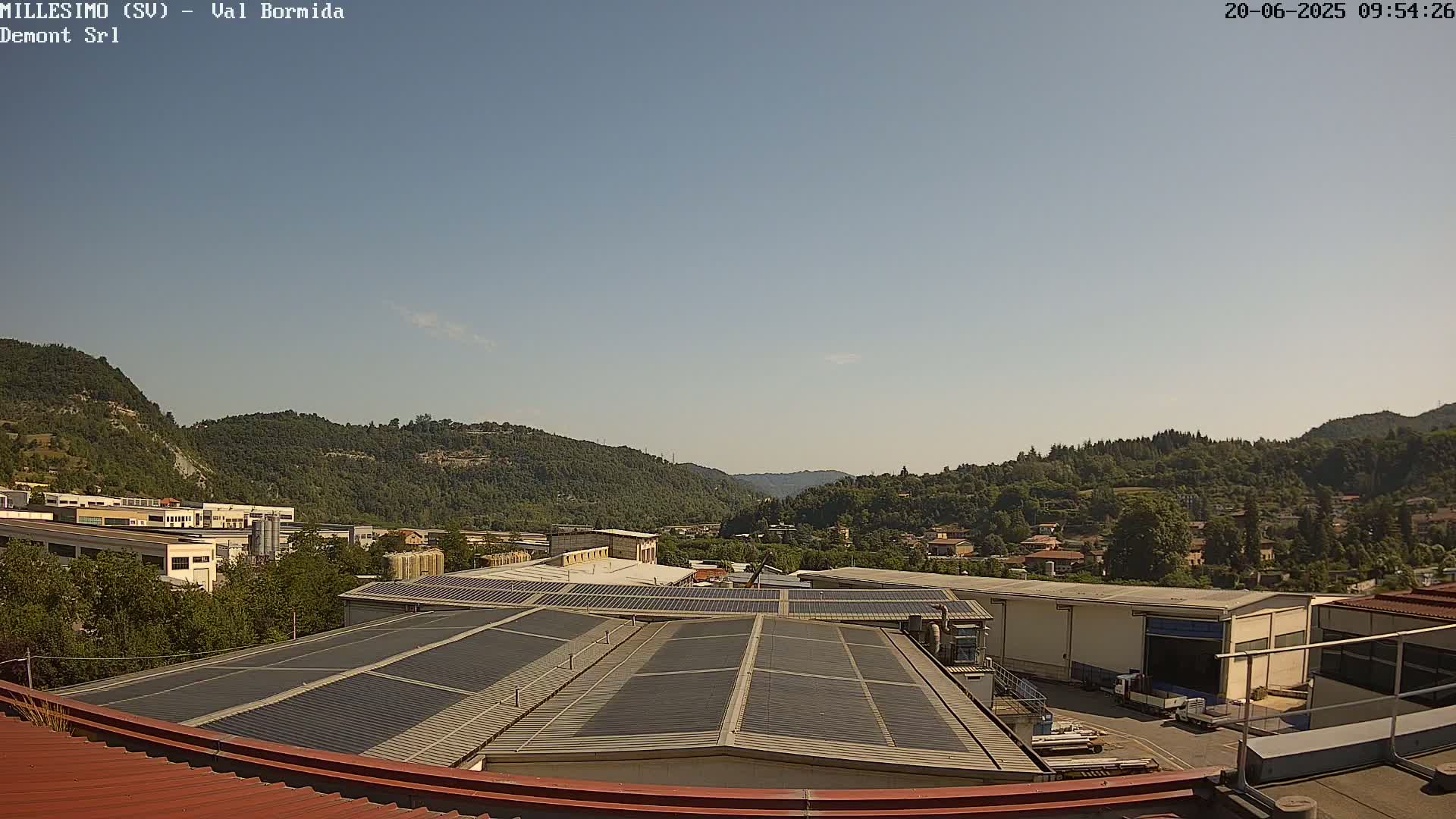 A view from a rooftop shows industrial buildings and a valley filled with green hills under a clear sky.