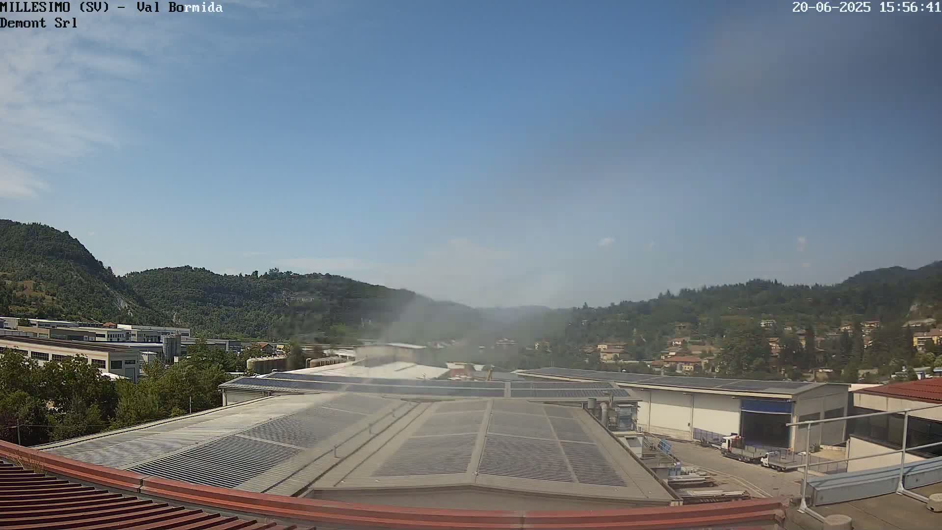 A hazy, sunny day overlooking an industrial area with solar panels on rooftops, nestled between green hills.
