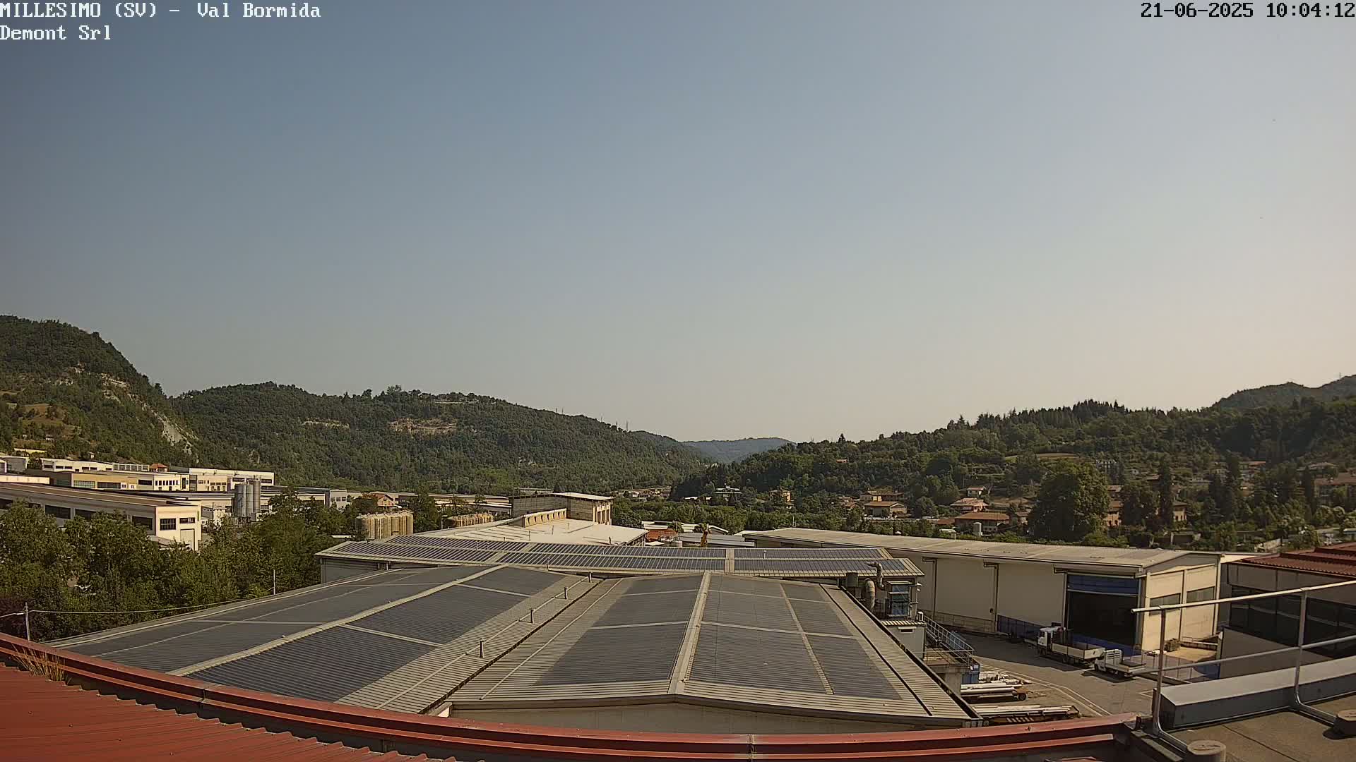 A clear, sunny day reveals a view of industrial buildings with solar panels on their roofs, nestled among green hills and a small town in the distance.