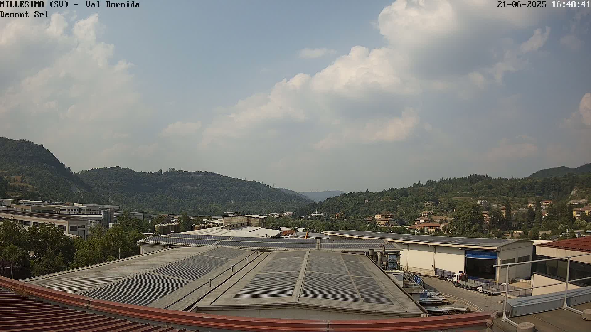 A partly cloudy sky overlooks industrial buildings with solar panels on their roofs, nestled in a valley surrounded by green hills.