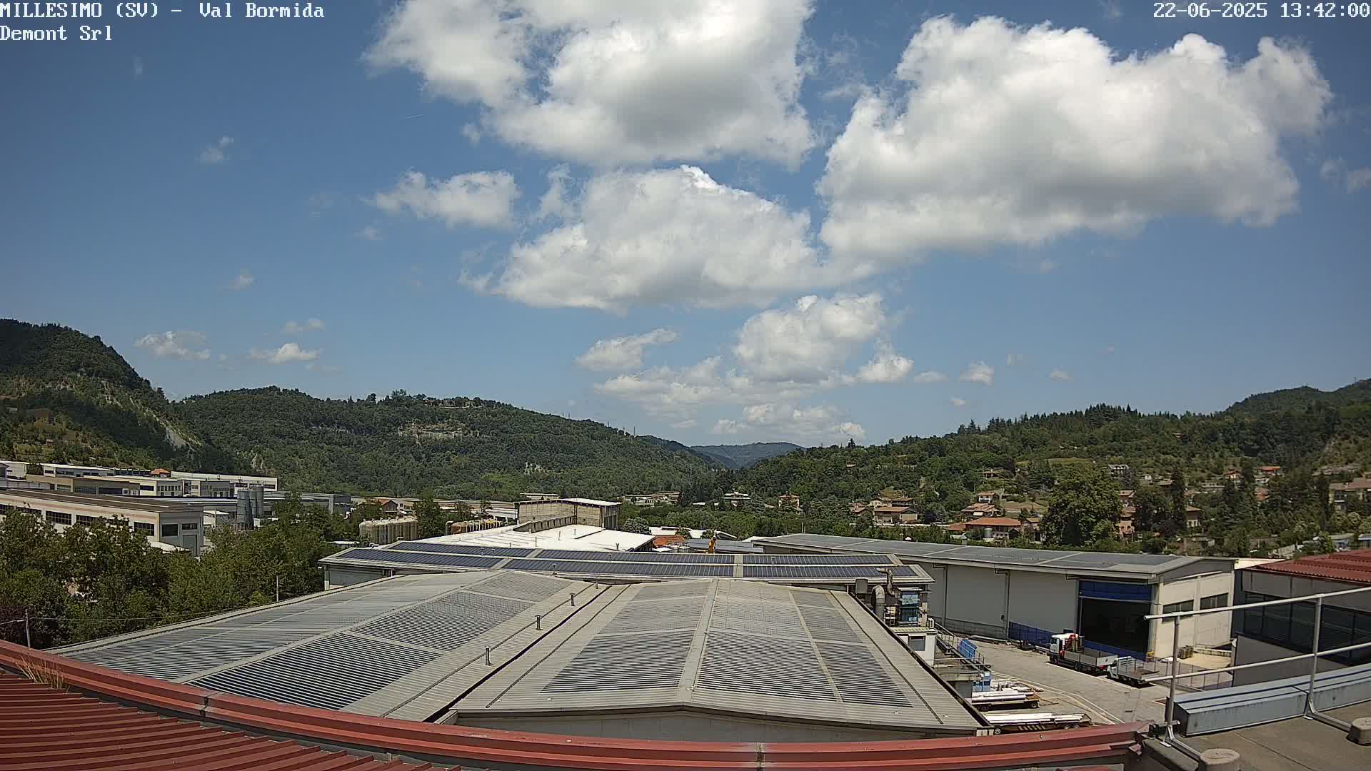 A partly cloudy sky overlooks a valley of green hills and industrial buildings with solar panels on their roofs.