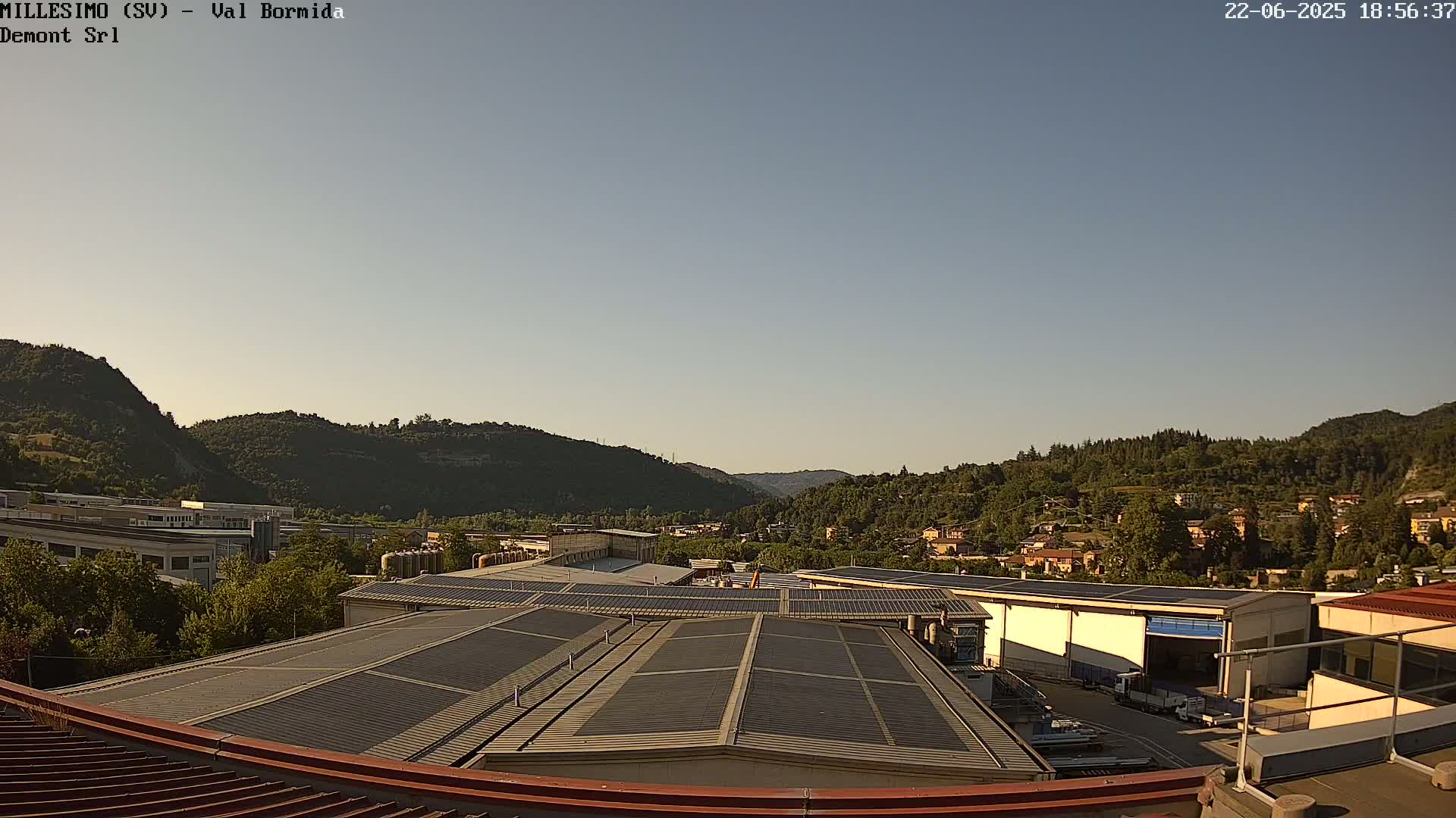 A view from a rooftop shows industrial buildings with solar panels, nestled amongst green hills under a clear, light-blue sky.