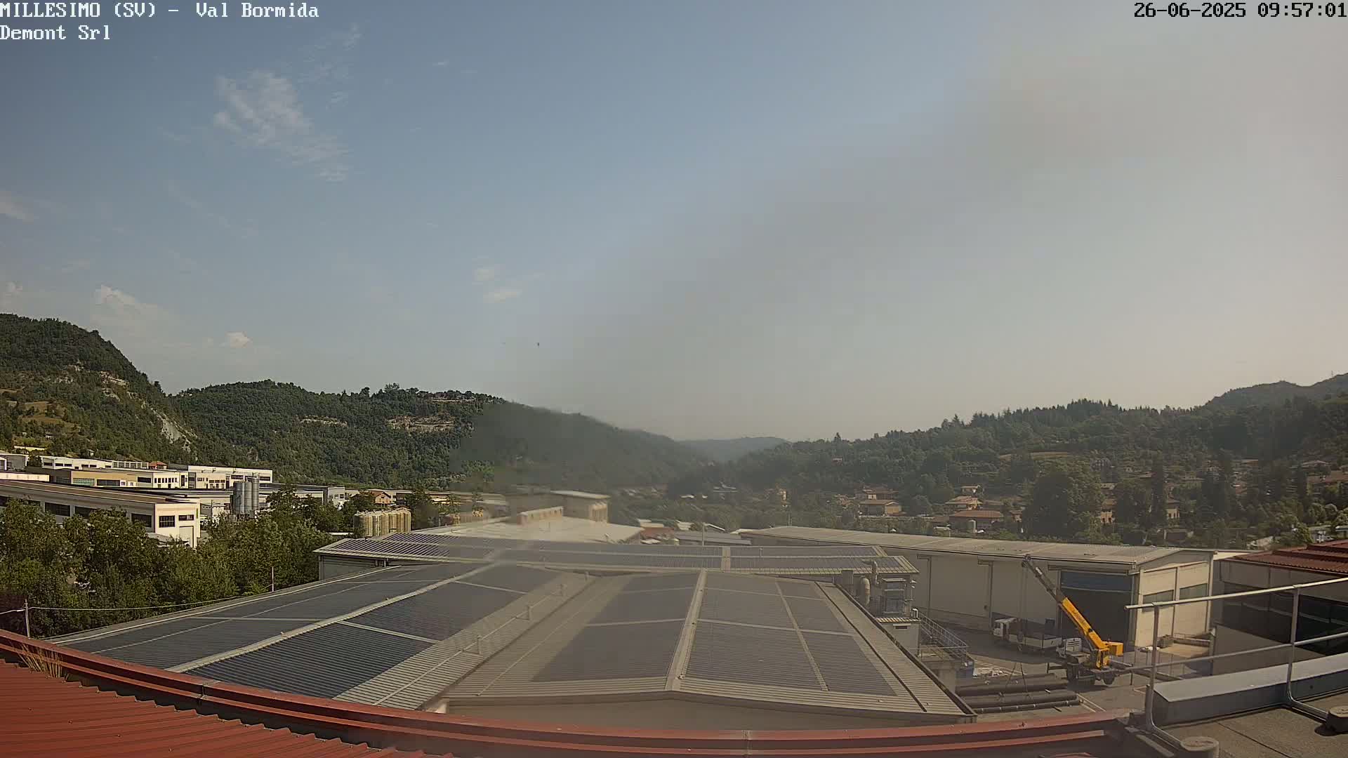 A hazy, sunny day reveals a view of industrial buildings and a small town nestled in a valley surrounded by tree-covered hills.