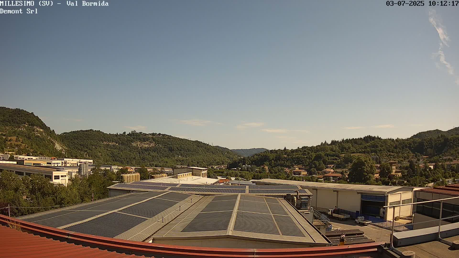 A clear, sunny day overlooks industrial buildings with solar panels on their roofs, nestled in a valley surrounded by green hills.