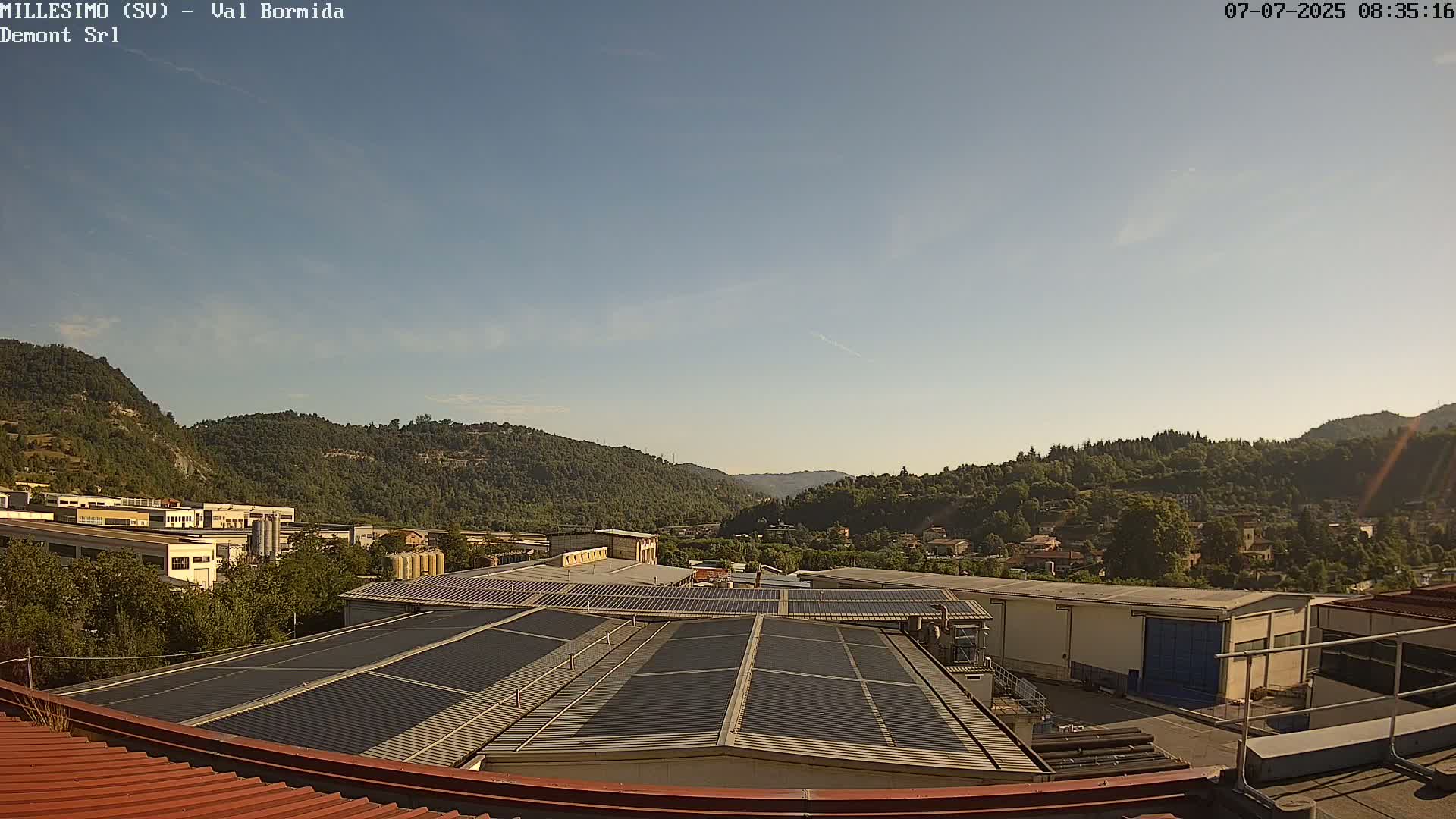 A view from a rooftop shows industrial buildings and a valley of green hills under a clear, bright sky.