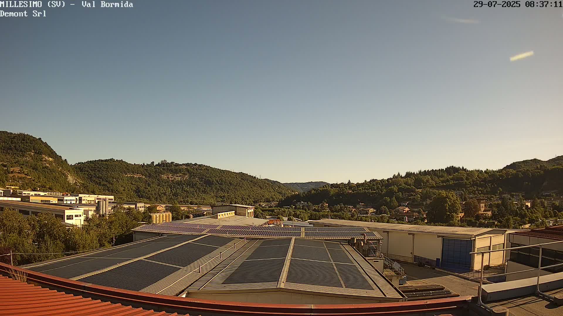 A clear, sunny day reveals a valley of green hills and trees, with industrial buildings and solar panels on rooftops in the foreground.