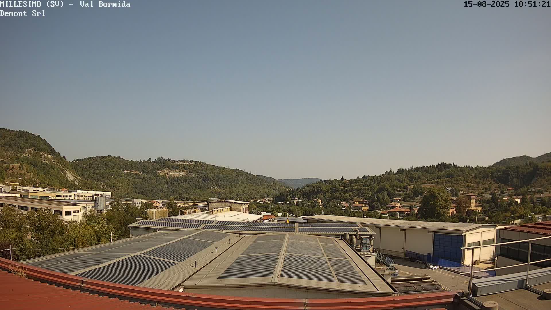 A clear, sunny day reveals a valley of green hills and trees, with industrial buildings and solar panels on rooftops in the foreground.
