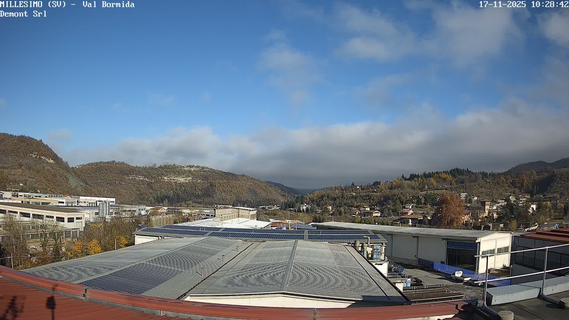 An elevated view captures an industrial area with solar-paneled roofs and autumn-tinged trees, transitioning to a town nestled among distant hills and a valley, all under a partly cloudy sky.