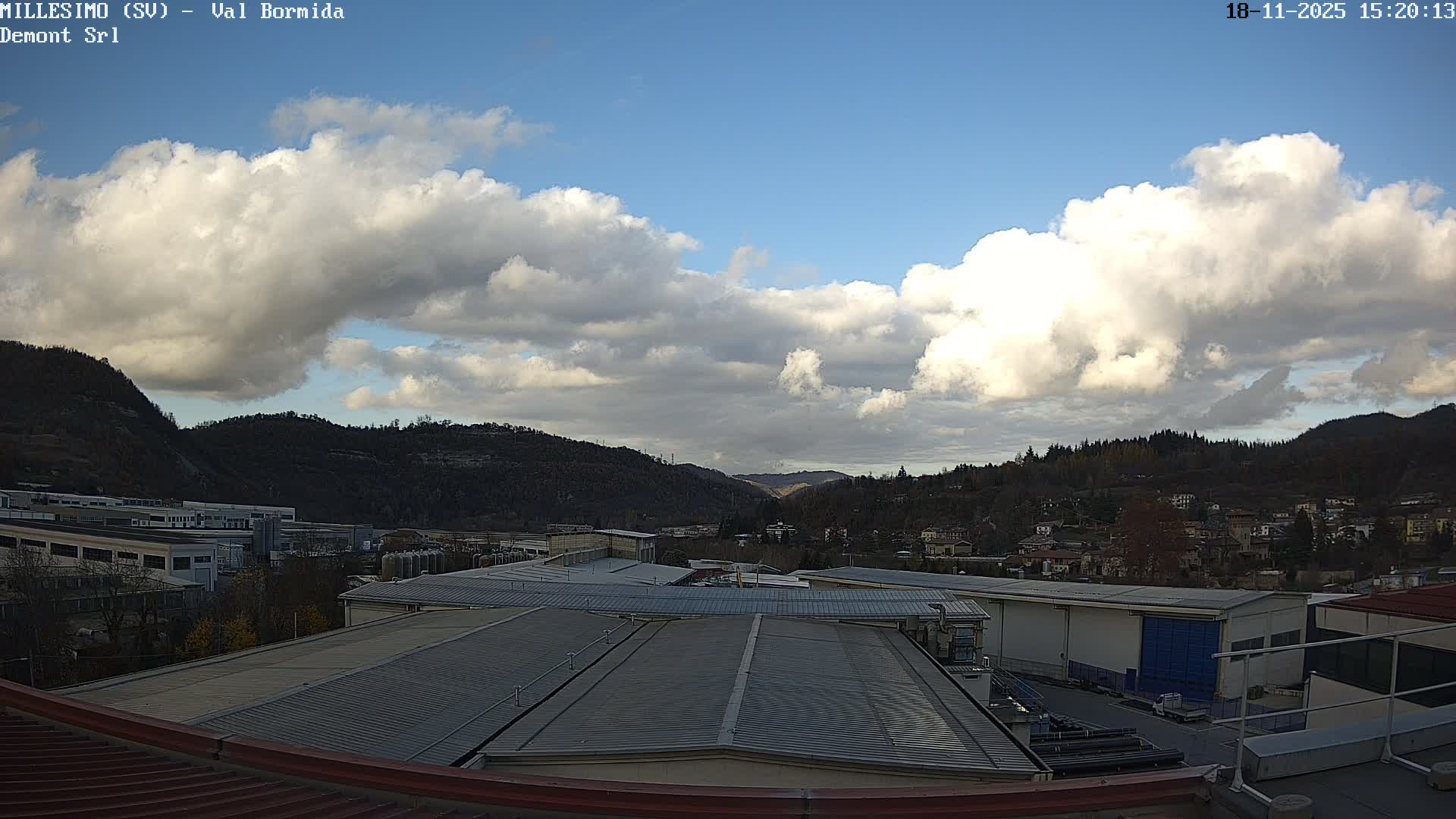 An elevated view captures an industrial area with solar-paneled roofs and autumn-tinged trees, transitioning to a town nestled among distant hills and a valley, all under a partly cloudy sky.