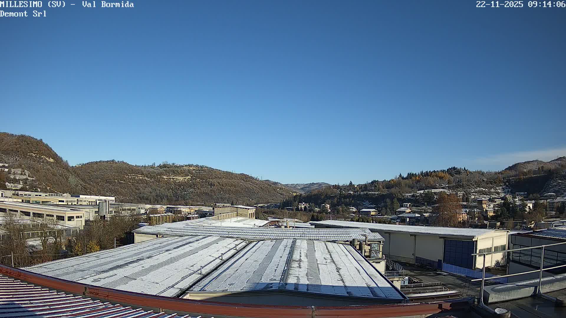 The image displays a winter scene under a clear, bright blue sky, with snow-dusted industrial rooftops in the foreground and a town nestled among snow-speckled hills and mountains in the background.