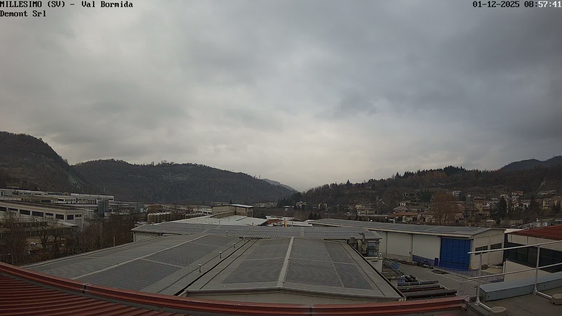 An overcast sky hangs over a valley featuring industrial buildings and residential areas, framed by tree-covered hills and mountains in the distance.