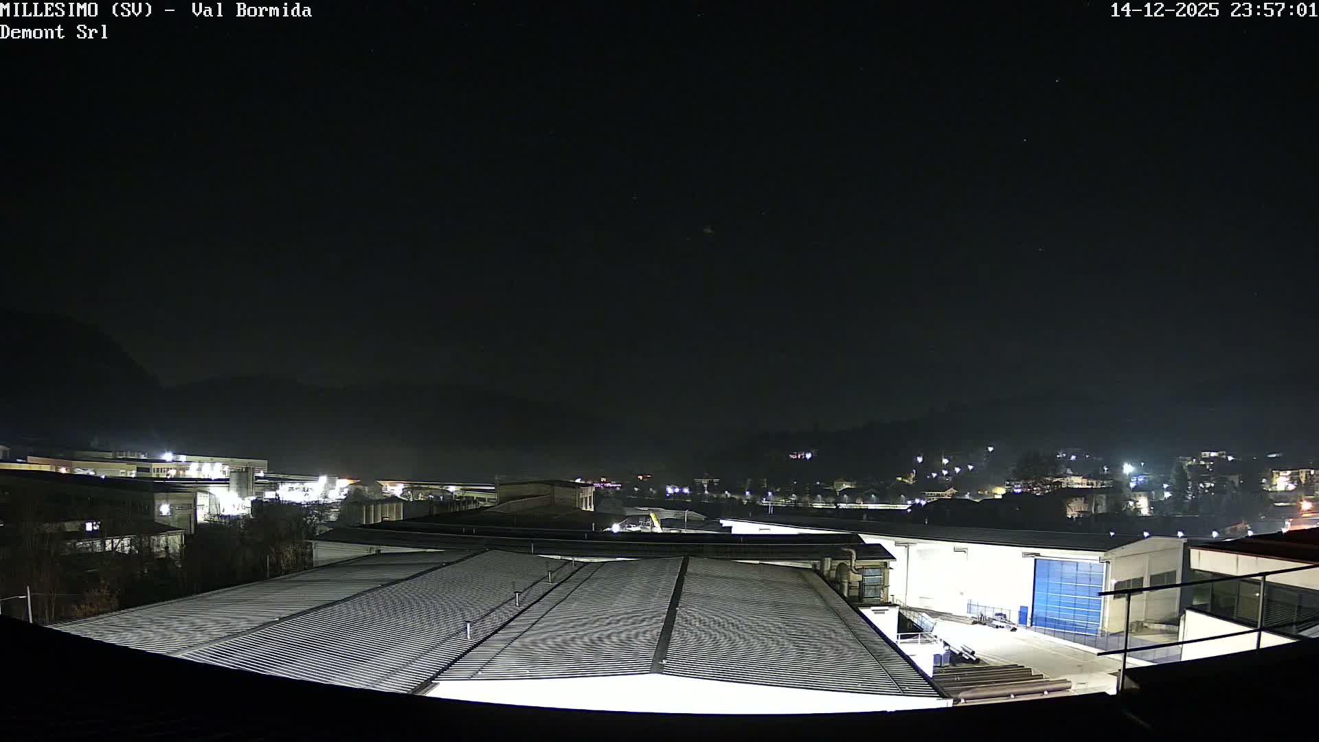 The image captures a clear night view of an industrial area and a brightly lit town situated in a valley, with dark mountain silhouettes against the starry and faintly clouded sky.