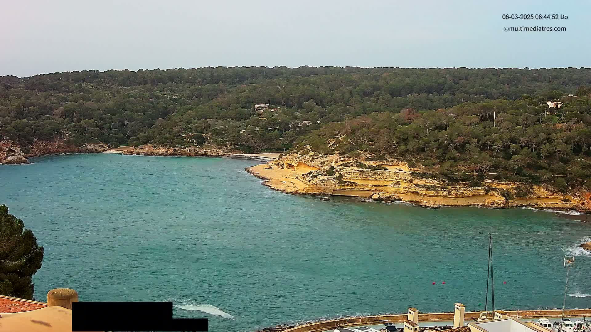 A tranquil cove with teal water, a sandy beach, and tan, eroded cliffs surrounded by lush green vegetation under an overcast sky.