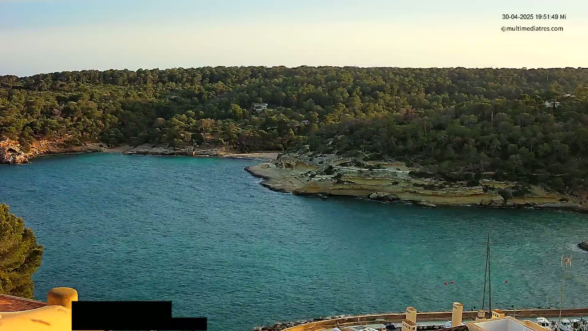 A tranquil cove with clear teal water is nestled amongst a verdant hillside under a calm, clear sky.