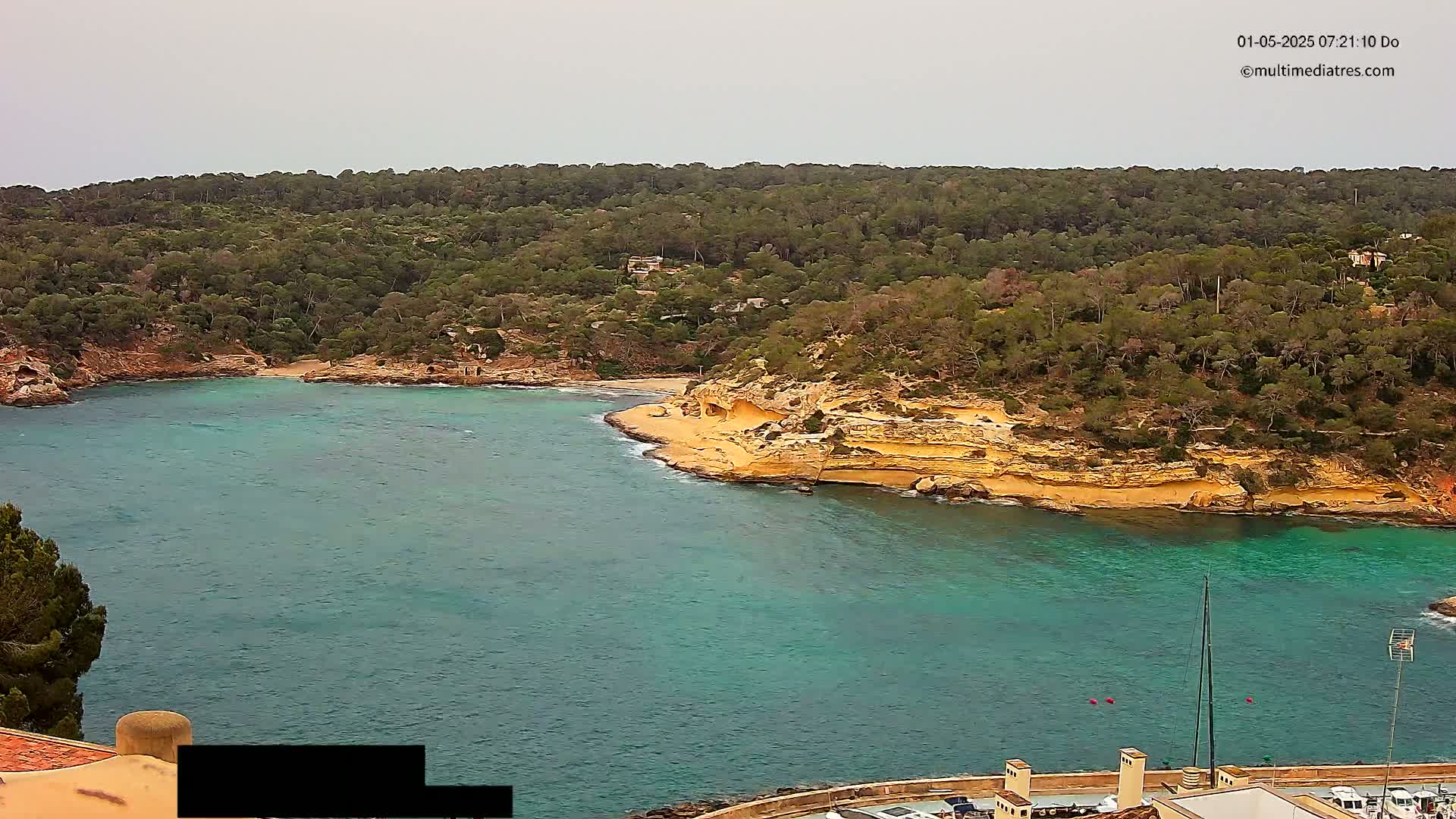 A tranquil cove with teal water is nestled within a verdant, hilly landscape under an overcast sky.