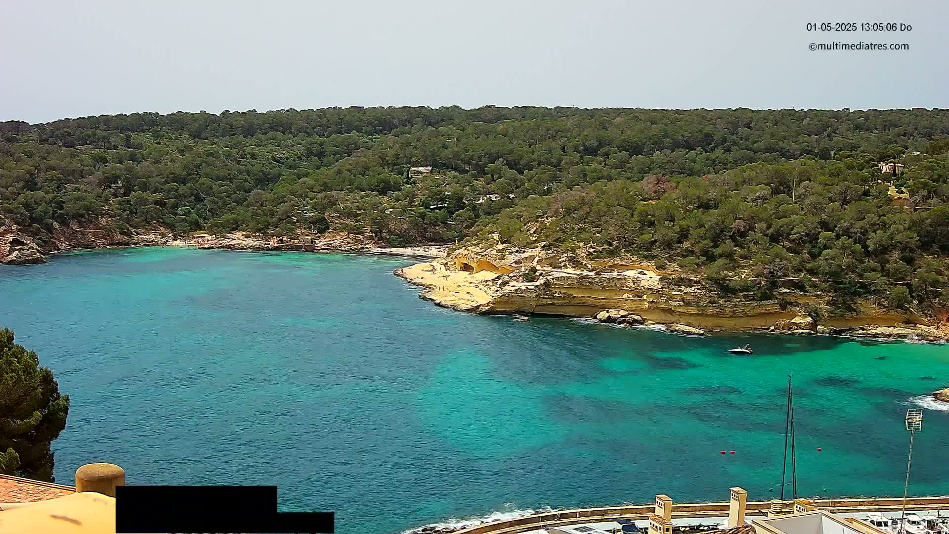 A tranquil cove with clear turquoise water is nestled amongst lush green hills under a partly cloudy sky.