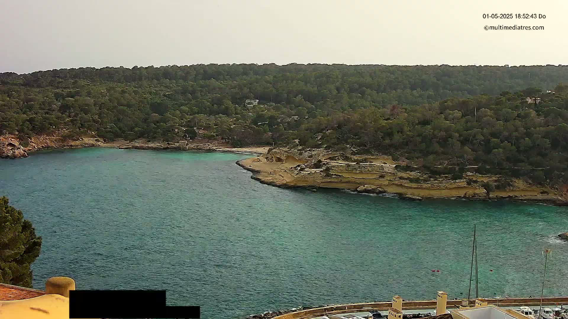 A tranquil cove with teal water is nestled between a sandy shore and lush green hills under a cloudy sky.