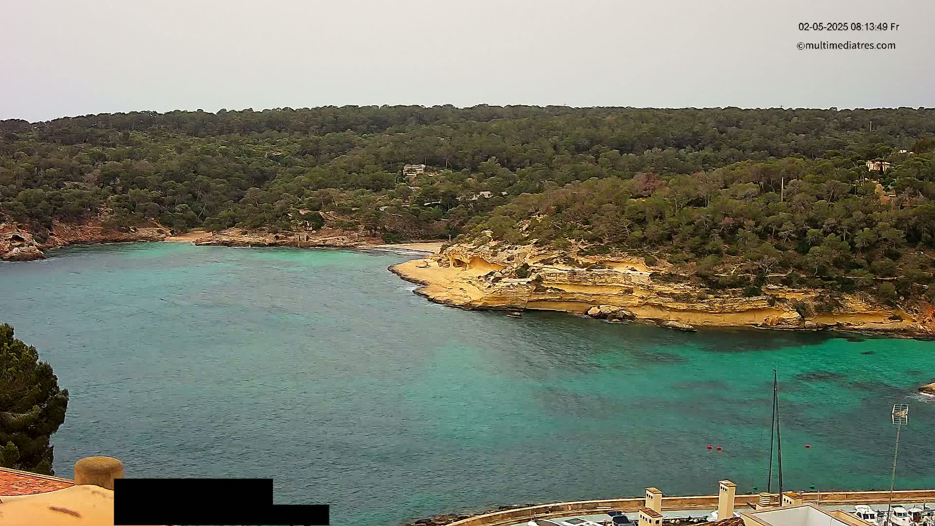 A secluded cove with a small sandy beach is surrounded by turquoise water and a lush, green hillside under an overcast sky.