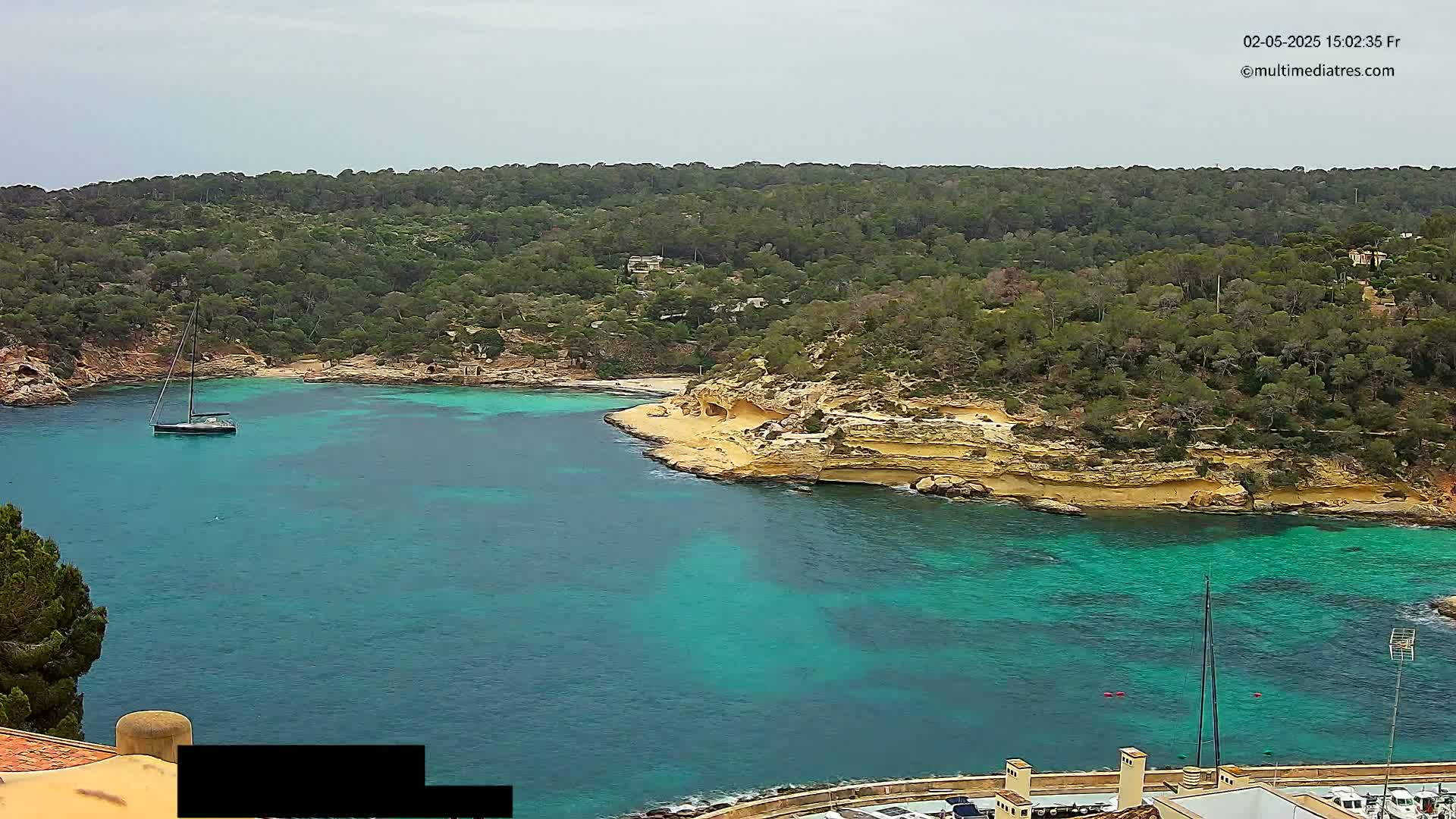A sailboat is anchored in a calm, turquoise bay surrounded by a lush, green hillside under an overcast sky.