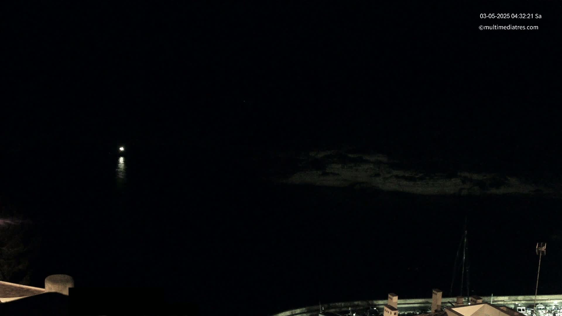 A dark, nighttime scene shows a body of water with a faint light in the distance, a dark shoreline, and some structures visible at the bottom of the frame.