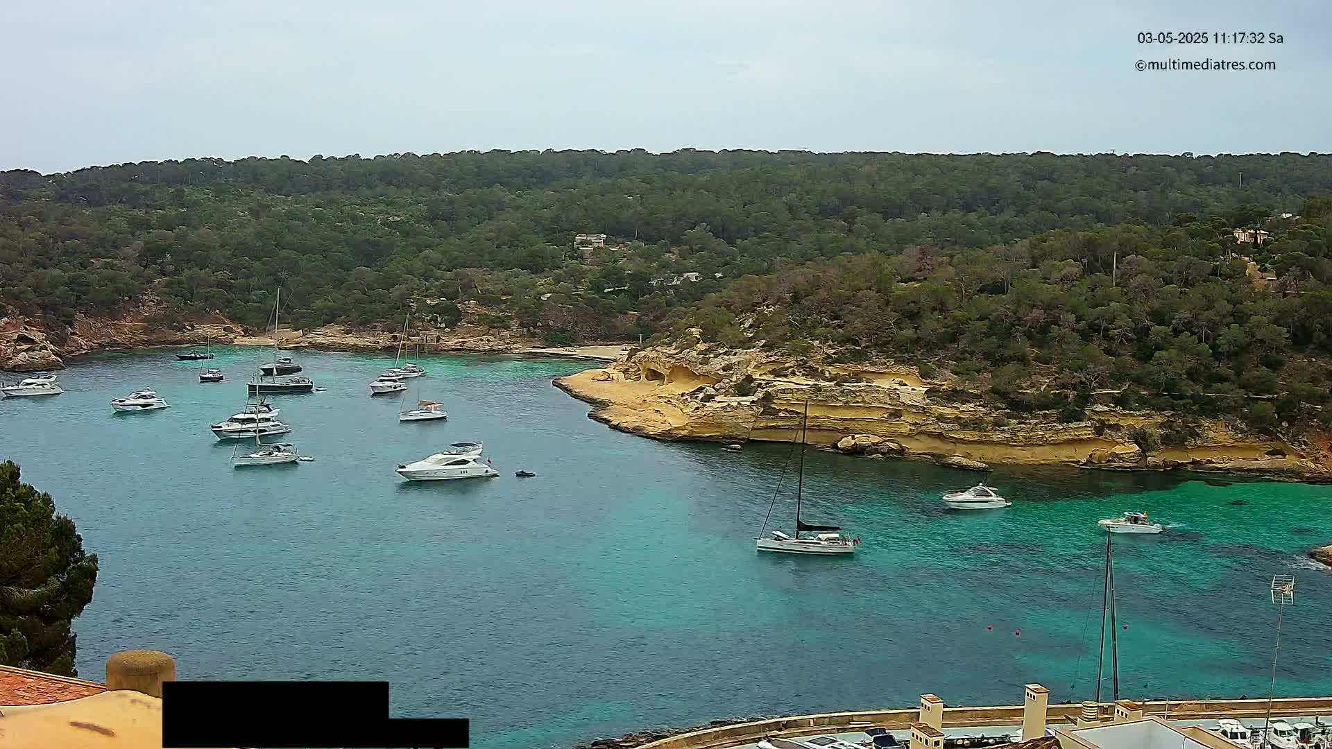 A calm bay with numerous boats and yachts is nestled within a cove surrounded by lush green hills under a mostly cloudy sky.