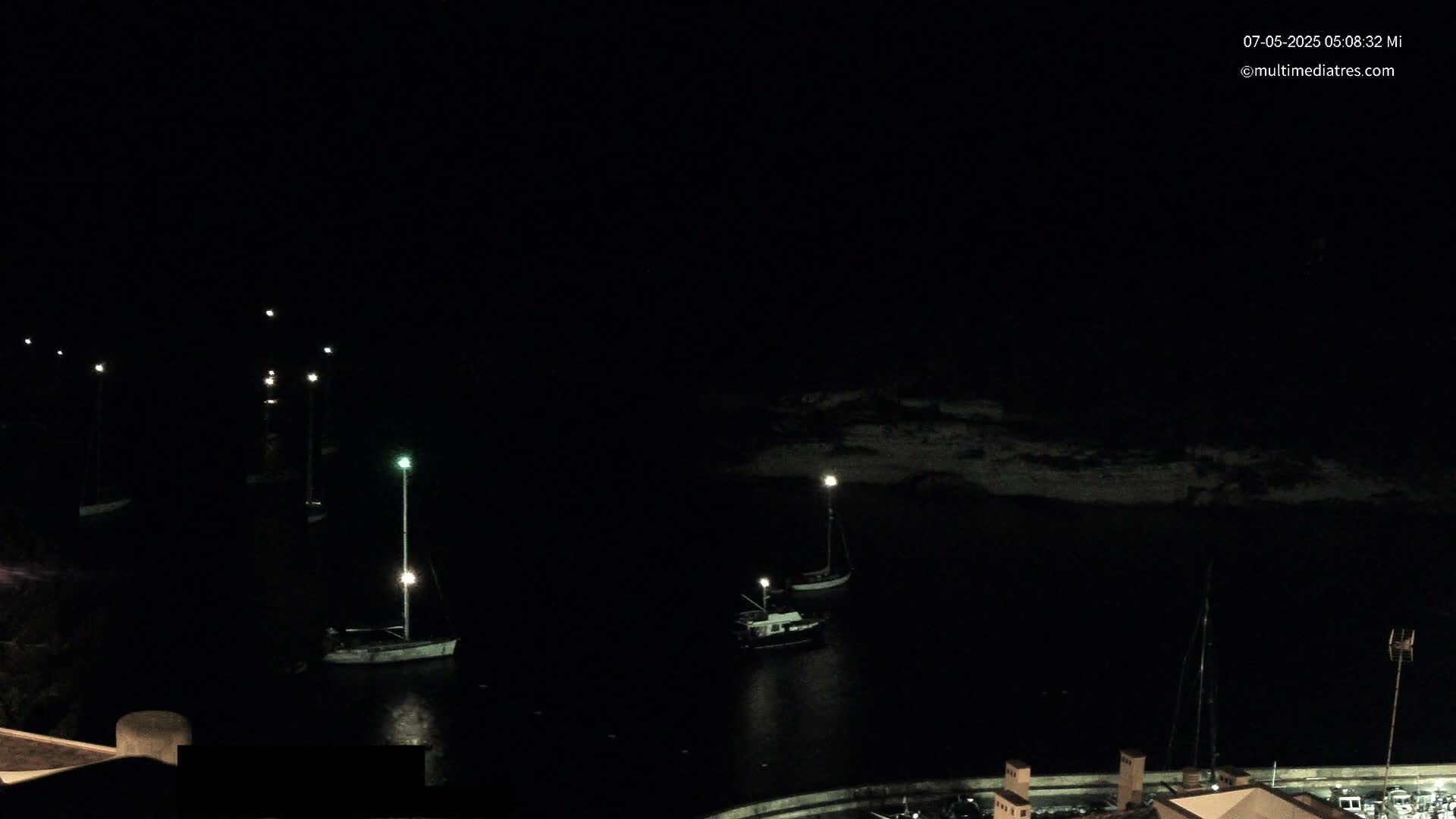 Several sailboats are moored in a dark harbor at night, illuminated by scattered lights.