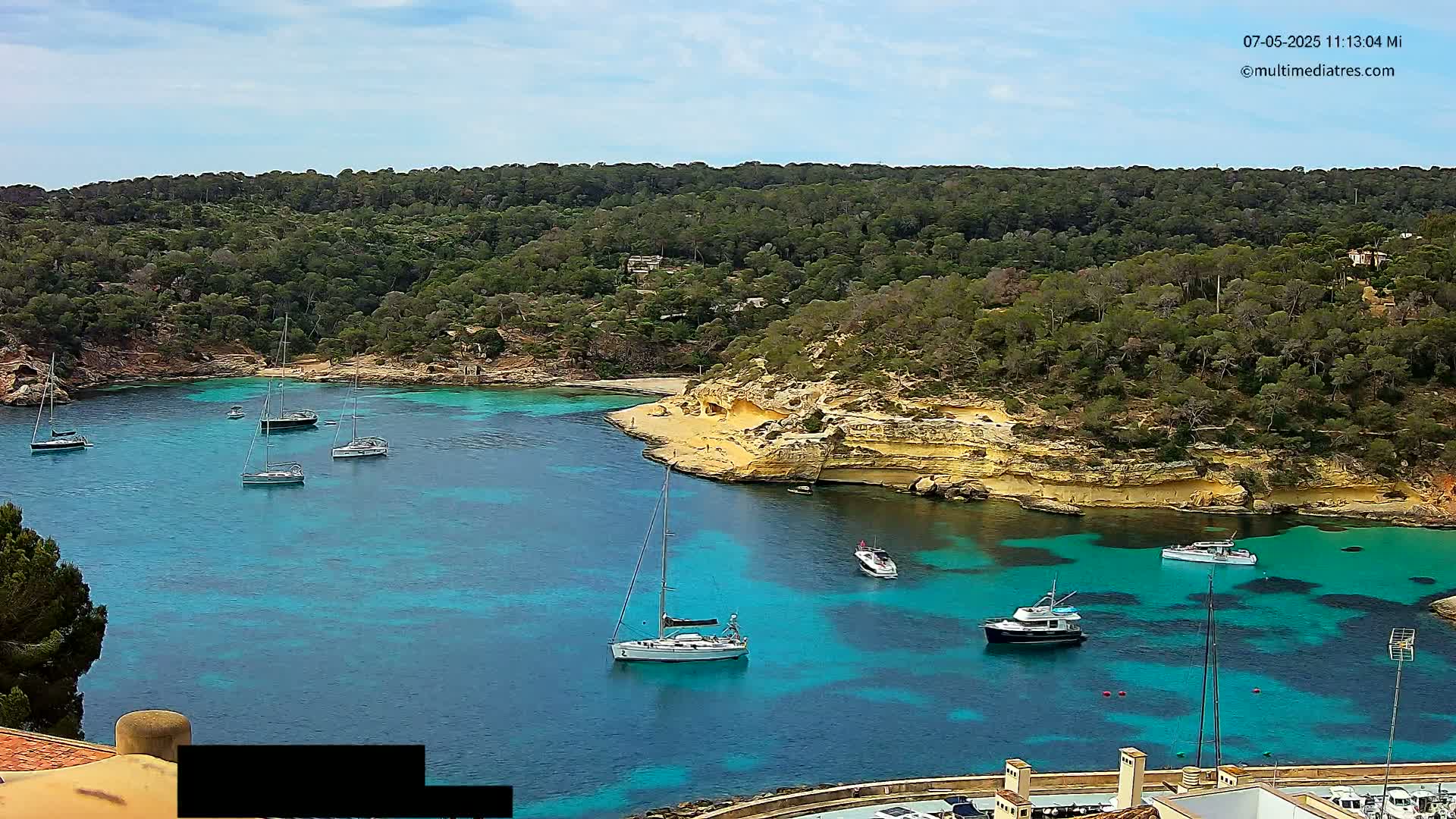 A tranquil cove with turquoise water, several sailboats and motorboats, and a sandy beach nestled within a lush, green hillside under a partly cloudy sky.