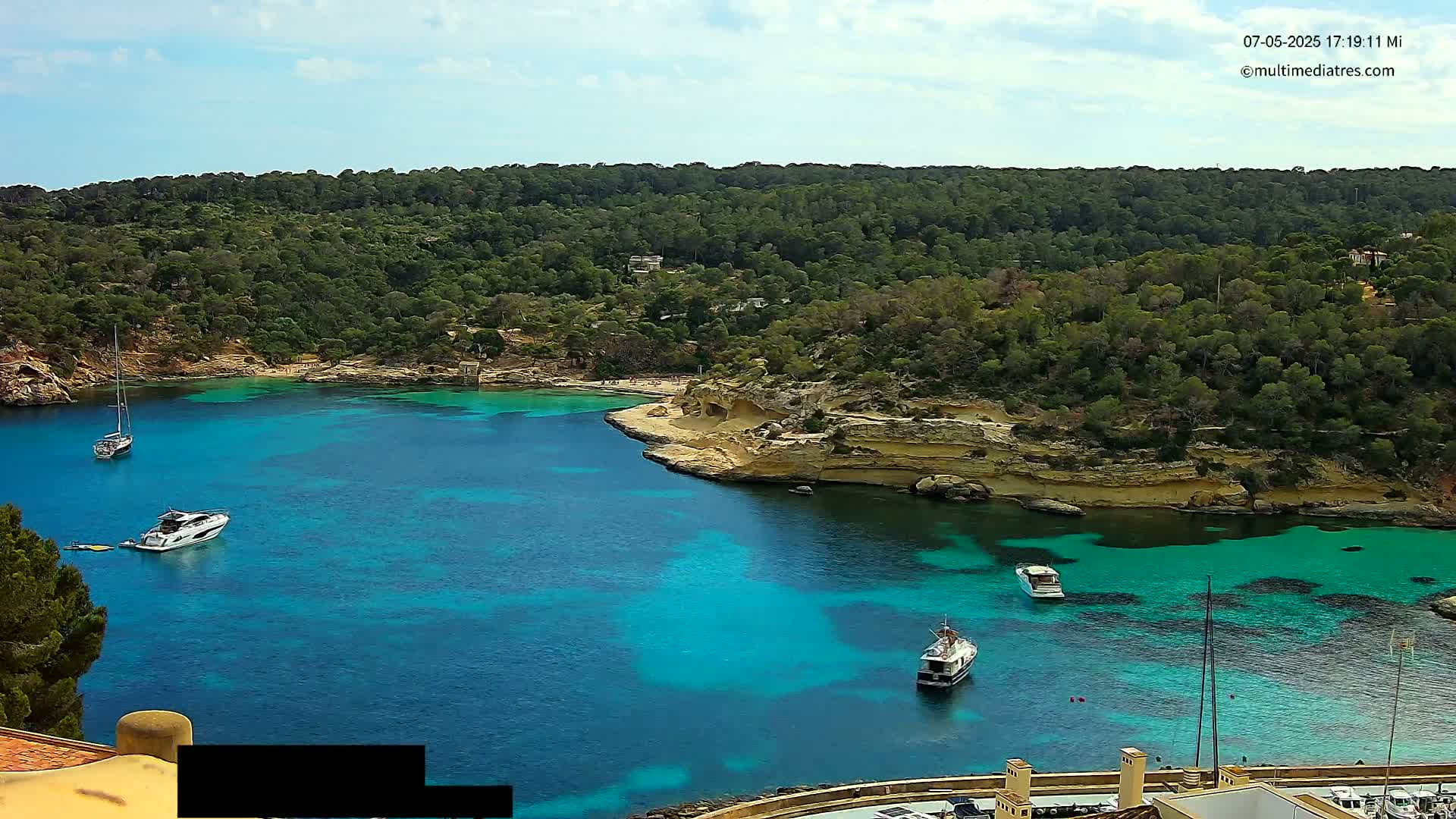 A tranquil cove with vibrant turquoise water is surrounded by lush green hills under a partly cloudy sky, with several boats moored in the calm waters.