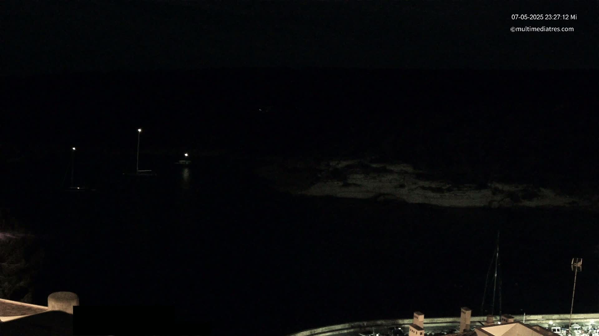 A dark nighttime view of a harbor with a few faintly lit boats and a shoreline visible under a clear, dark sky.