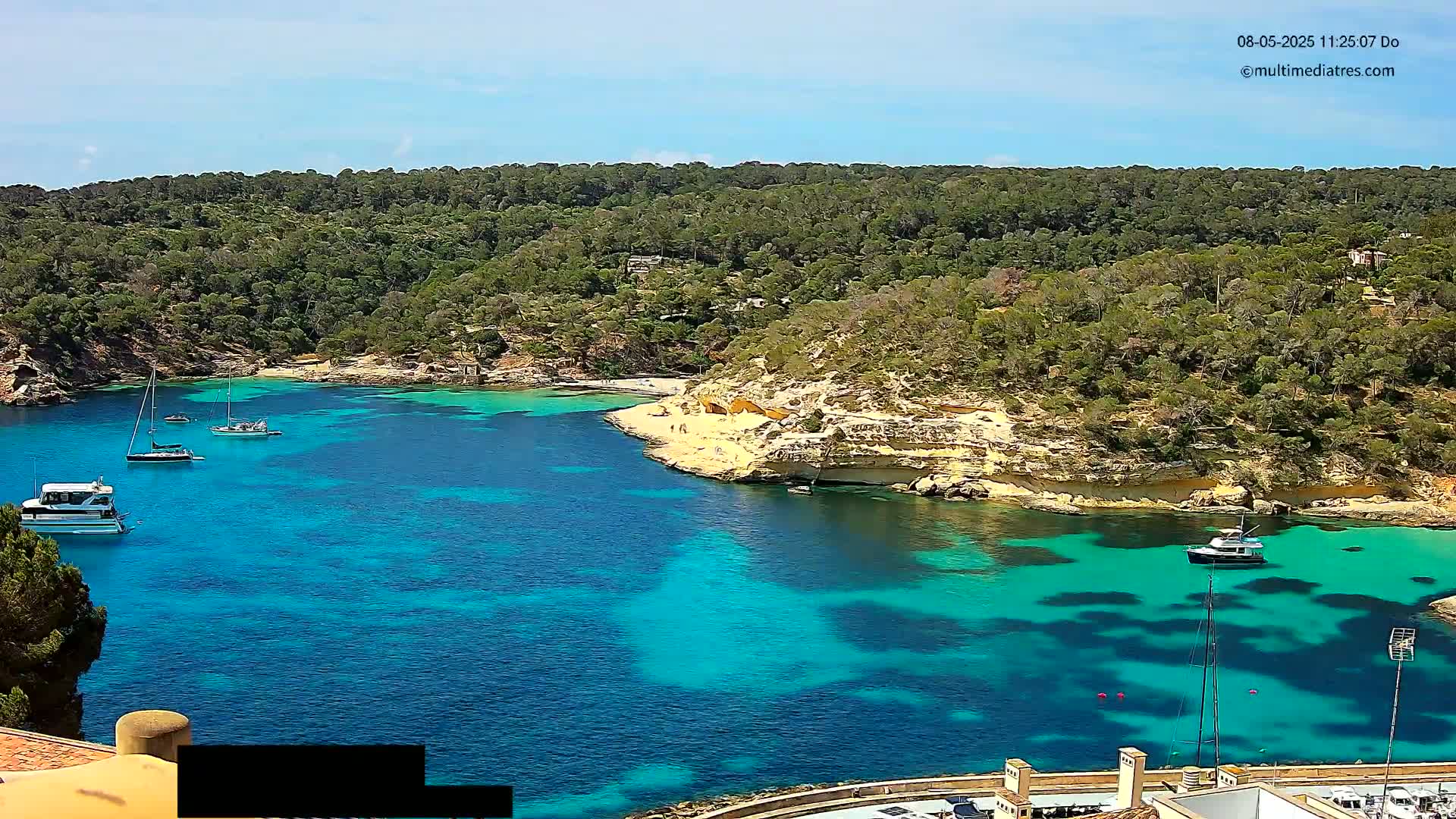 A secluded cove with bright turquoise water is surrounded by lush green hills under a clear, sunny sky, several boats are anchored in the bay.
