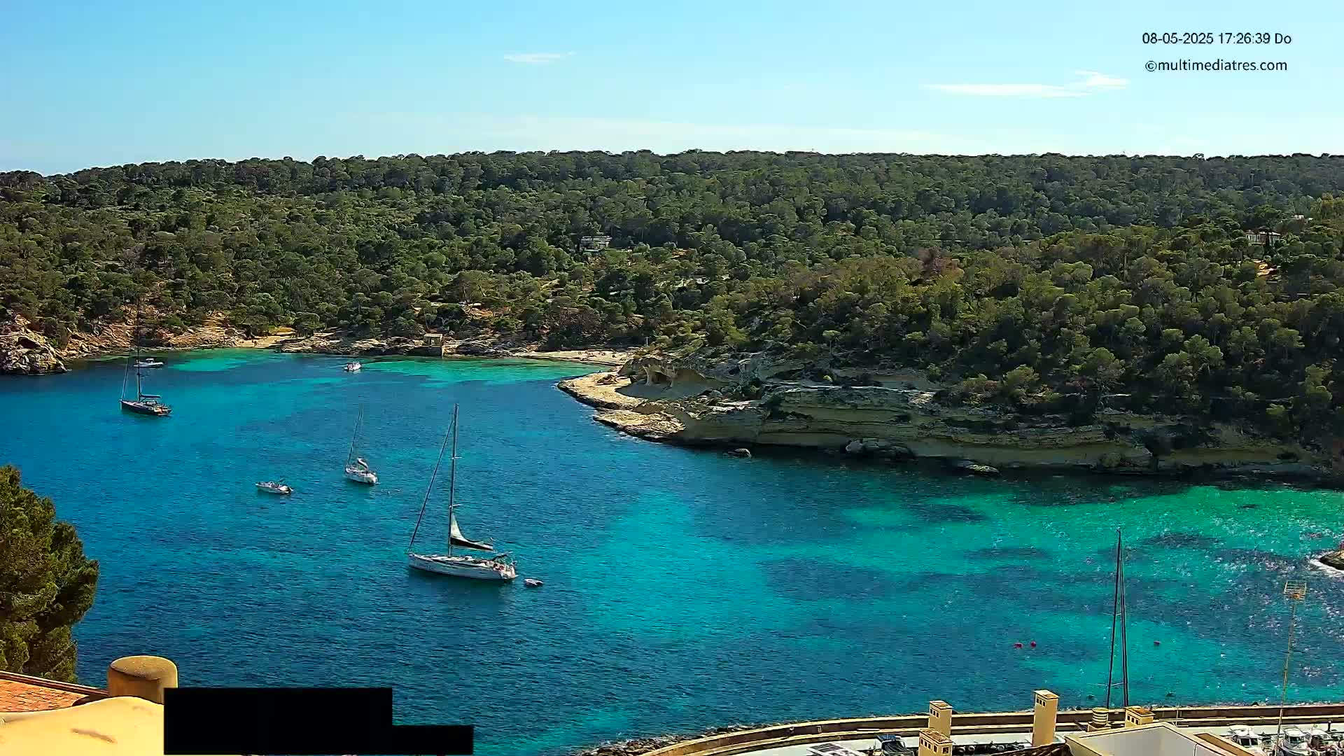 A secluded cove with turquoise water holds several sailboats, surrounded by a lush green hillside under a clear blue sky.