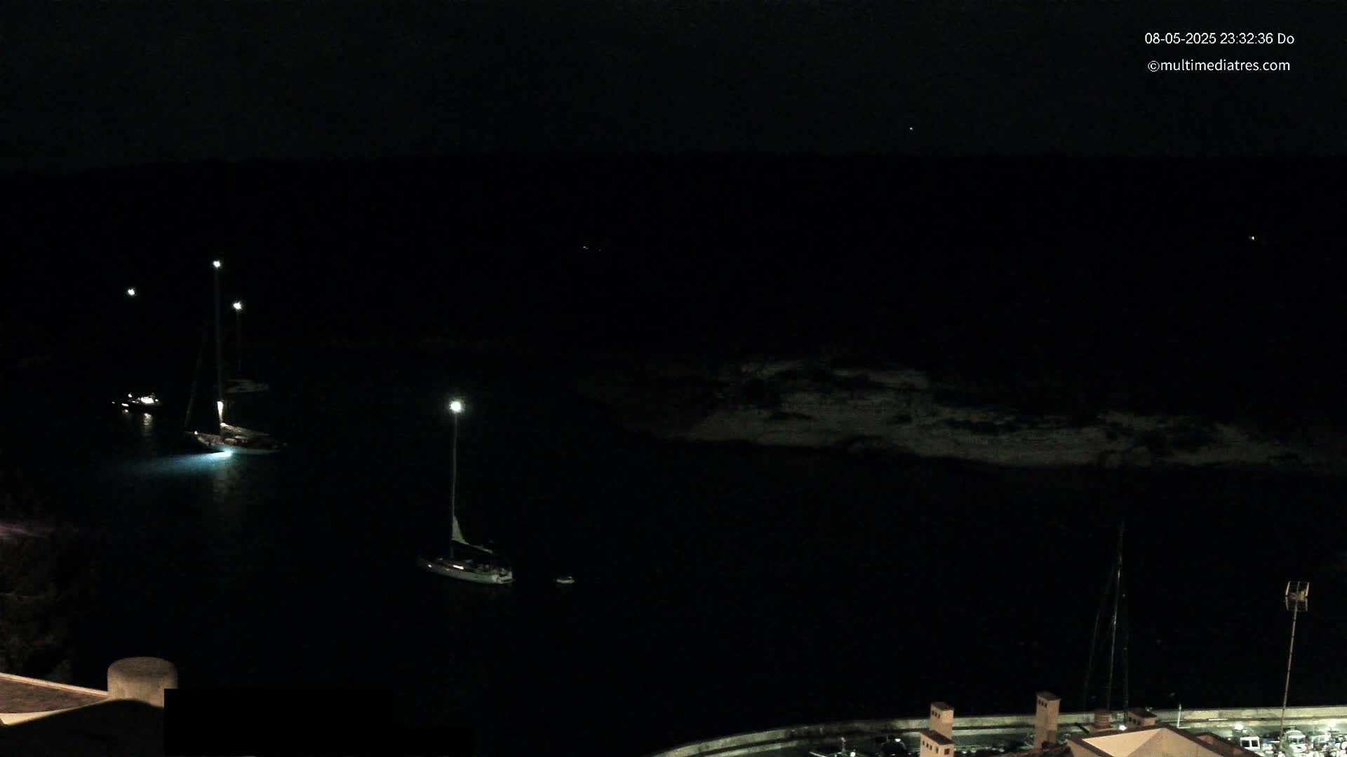 Several sailboats are moored in a dark, calm harbor at night, illuminated by lights on the docks.