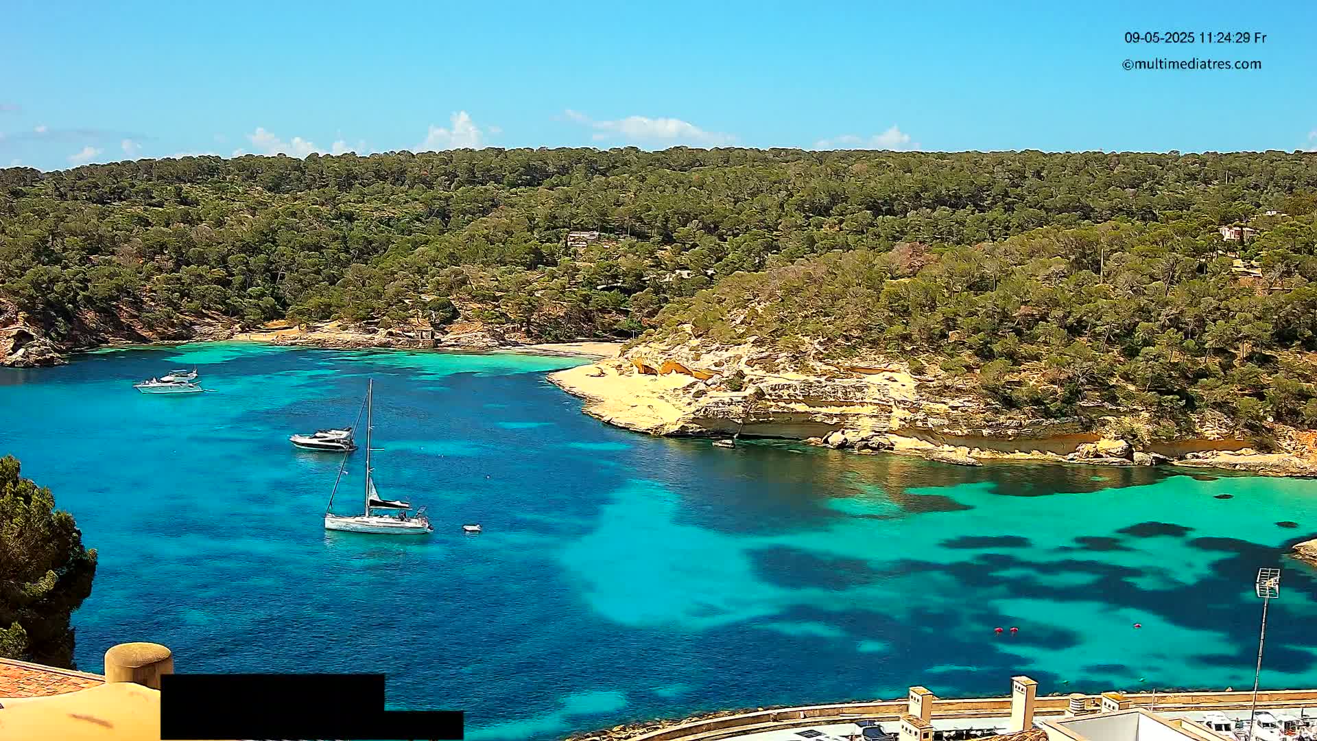 A secluded cove with turquoise water is surrounded by lush green hills under a bright sunny sky, with several boats anchored in the bay.