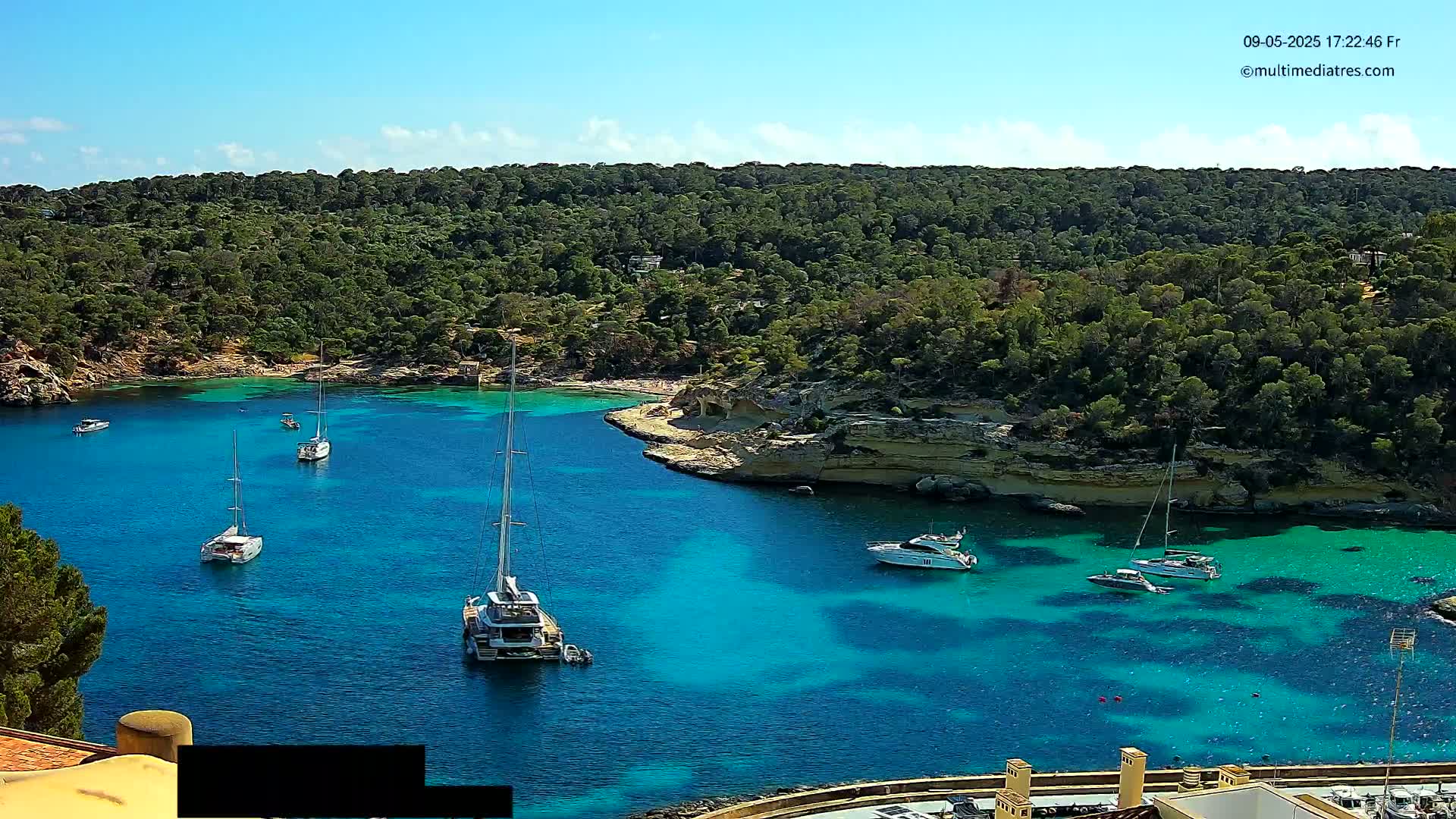 Several sailboats and motorboats are anchored in a clear, turquoise bay surrounded by lush green vegetation under a bright, sunny sky.