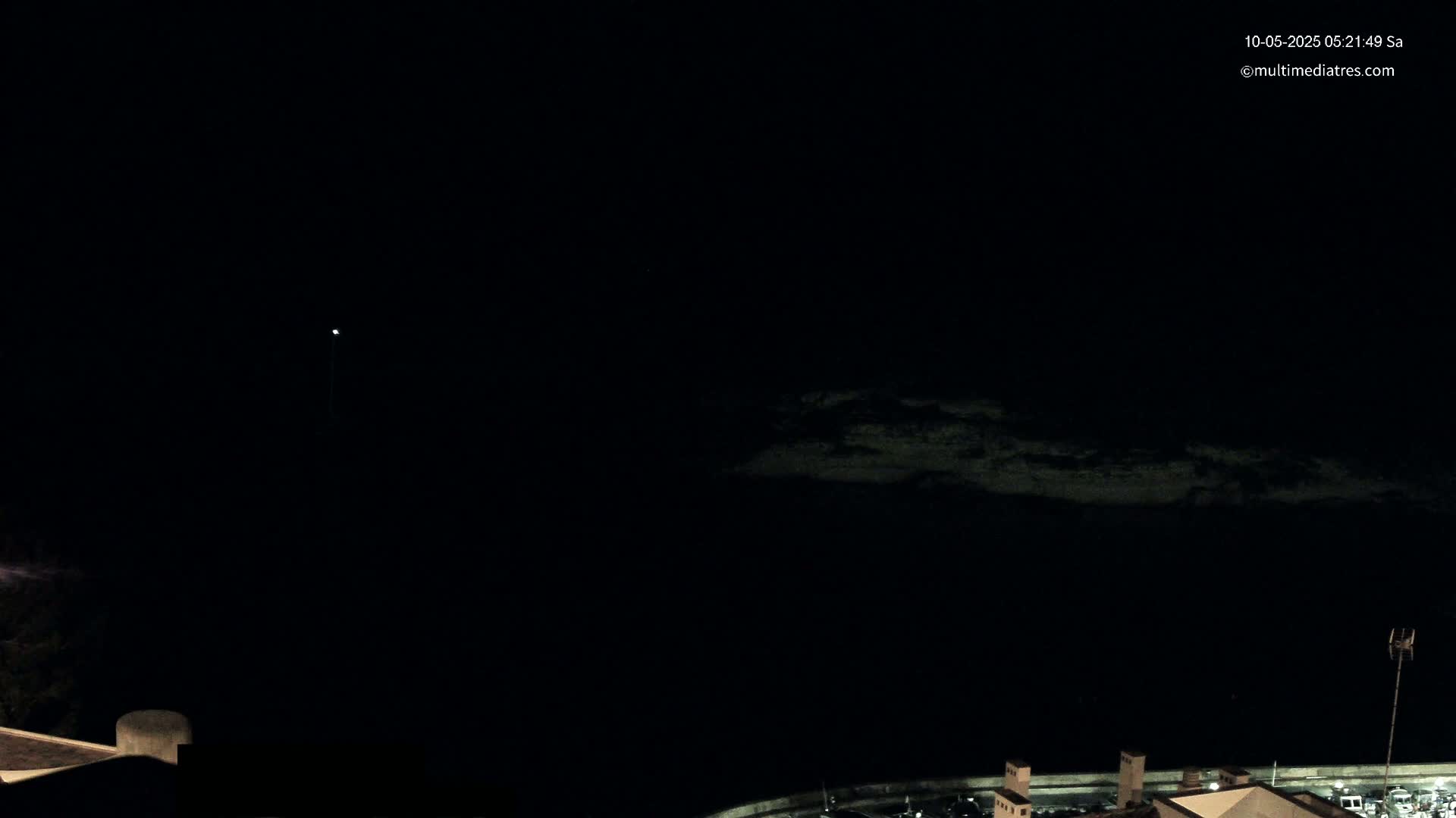 A dark nighttime scene shows a dimly lit body of water in the distance and a portion of a building in the foreground under a clear, dark sky.