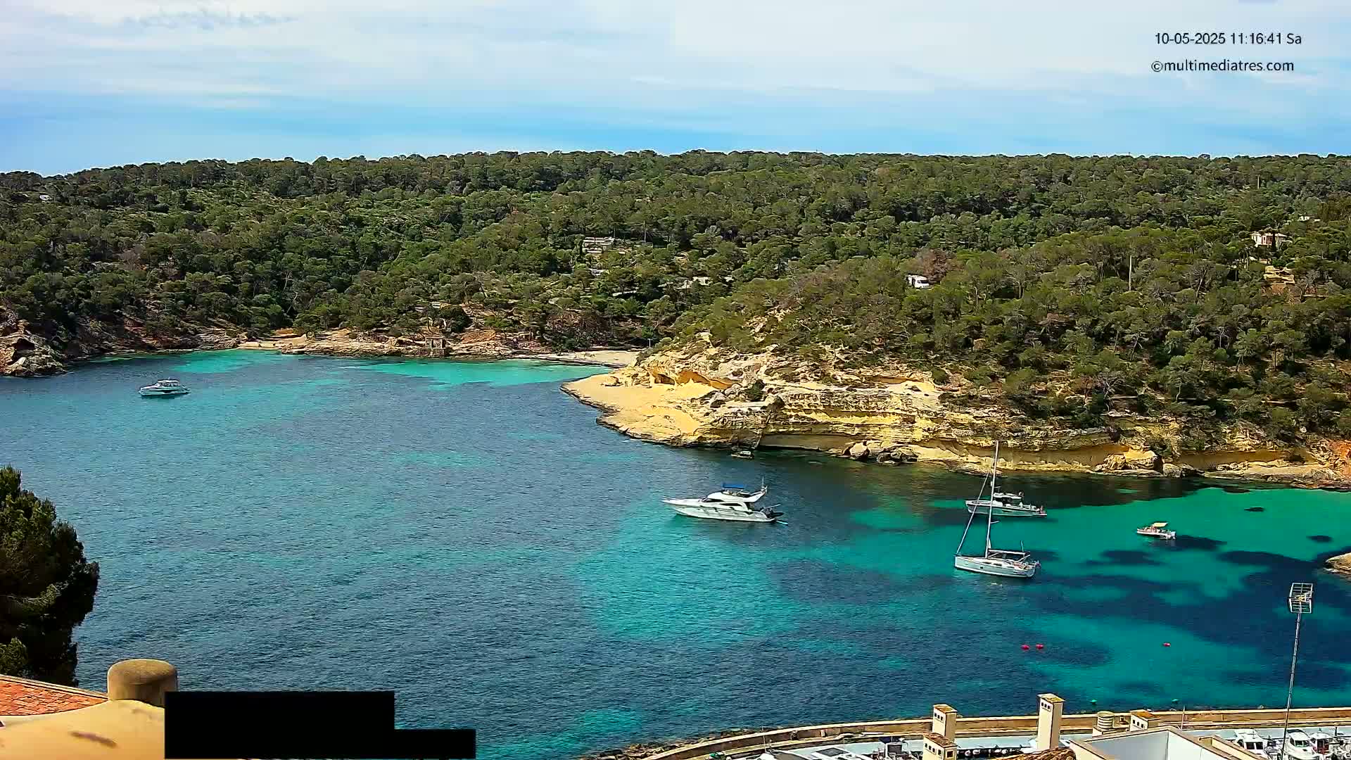 A secluded cove with clear, turquoise water is surrounded by lush green vegetation and rocky, light-brown cliffs under a mostly sunny sky.