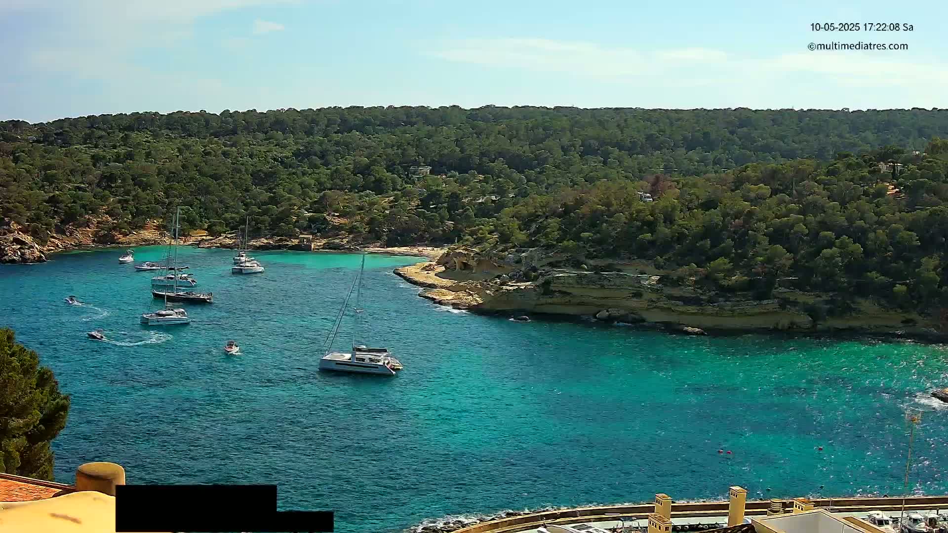 Several sailboats are anchored in a calm, clear turquoise cove surrounded by lush green hills under a sunny sky.