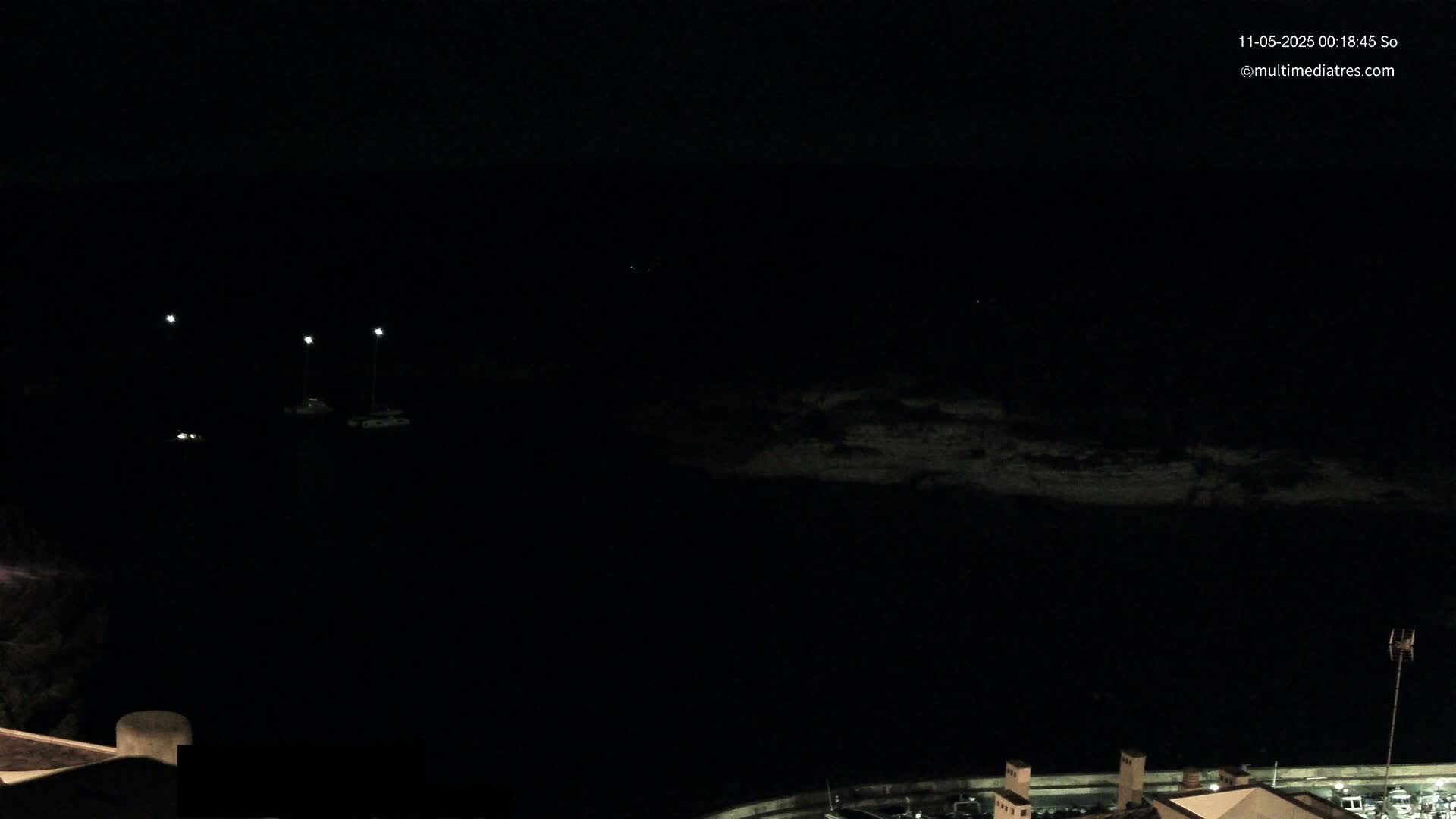 A nighttime, dark view of a body of water with faintly lit boats and a shoreline visible in the distance under a clear sky.
