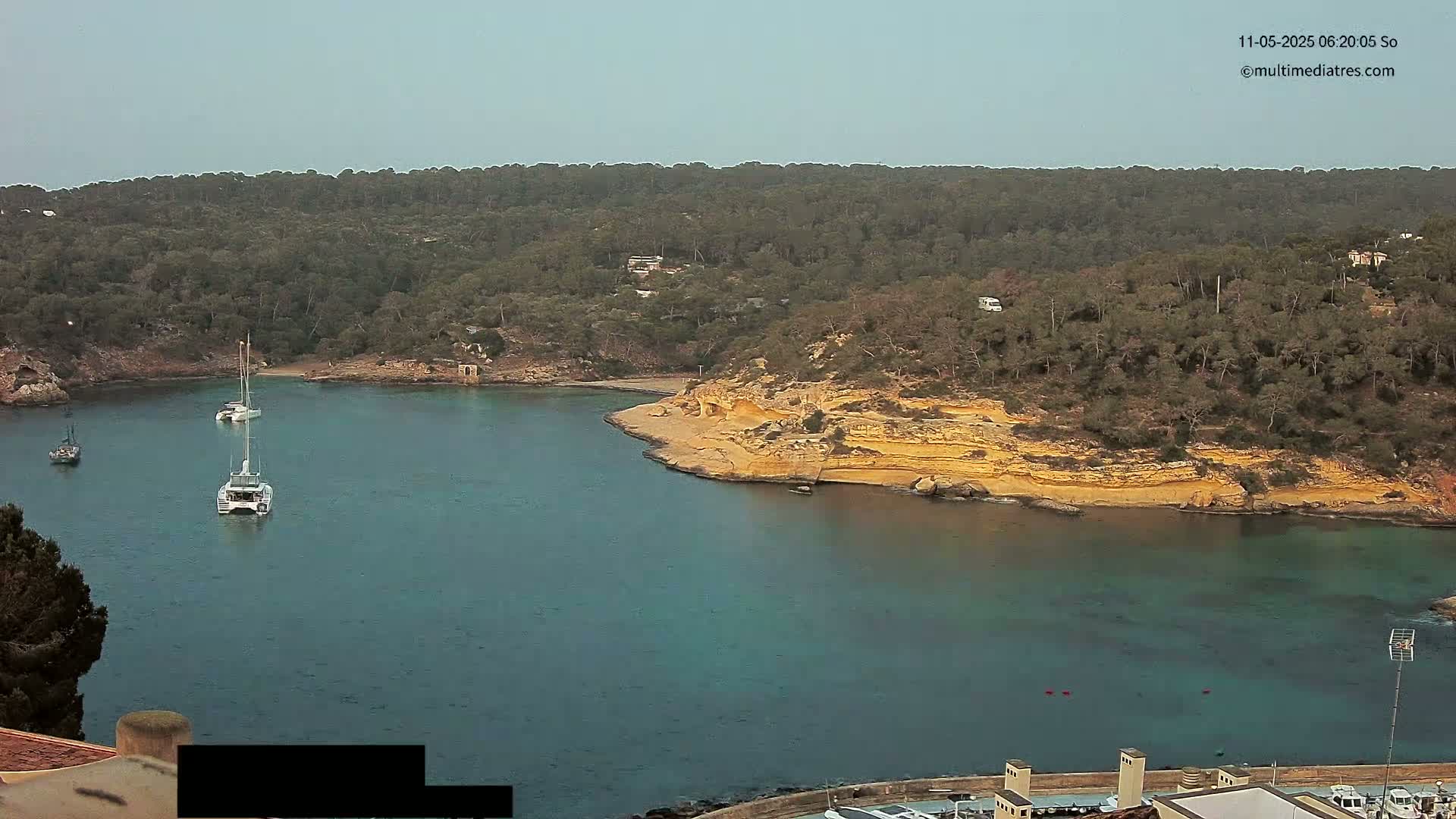 A calm bay, surrounded by a lush green hillside with a rocky, light-brown shoreline, contains several sailboats under a clear, light-grey sky.