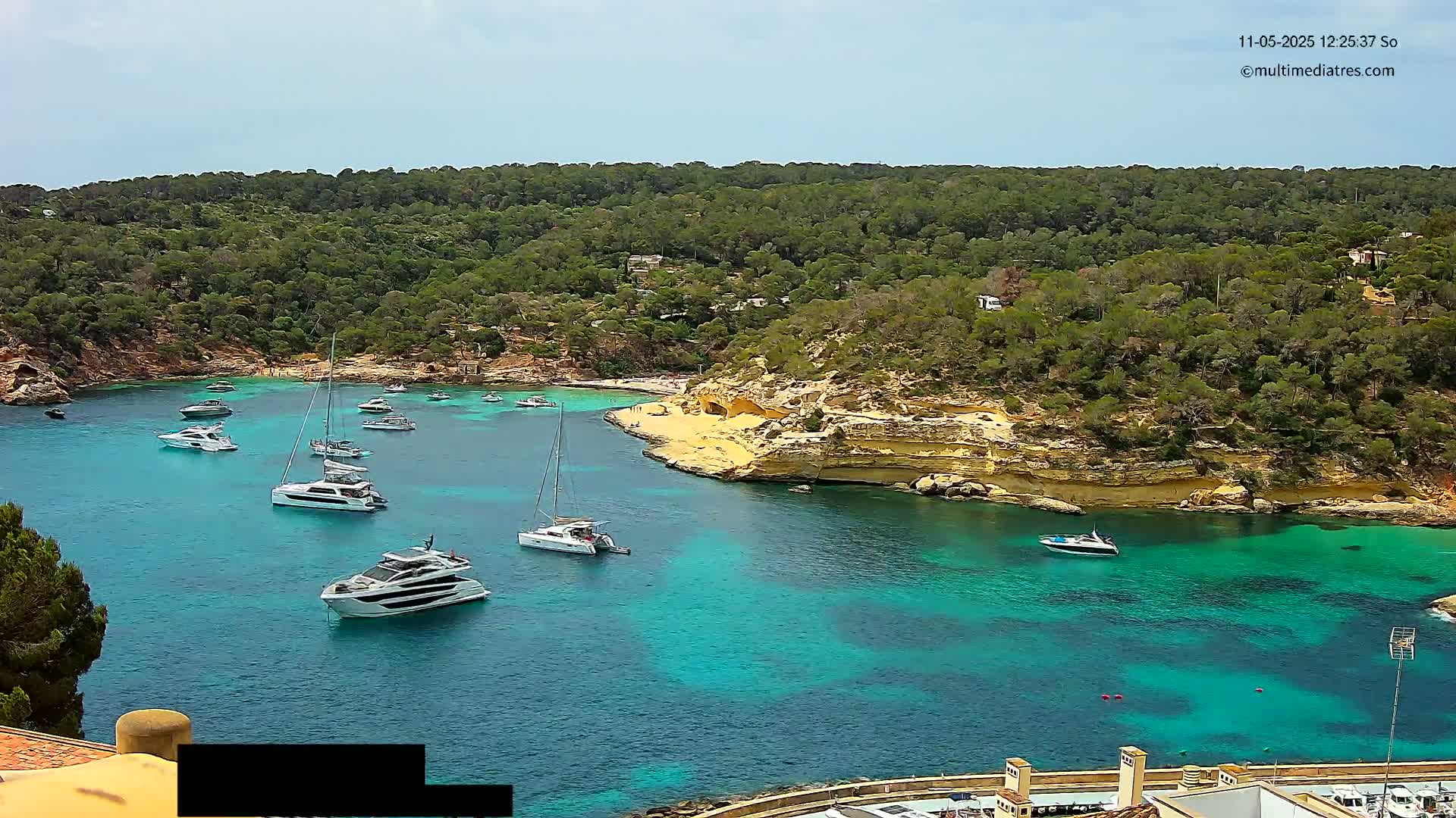 A tranquil turquoise cove, nestled within a lush green hillside, is dotted with several yachts and sailboats under a mostly sunny sky.