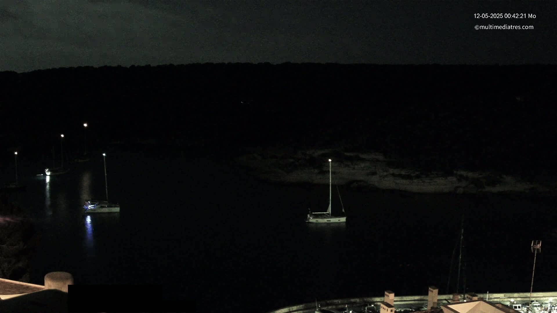 Several sailboats are moored in a dark, calm harbor at night, illuminated by faint lights on shore.