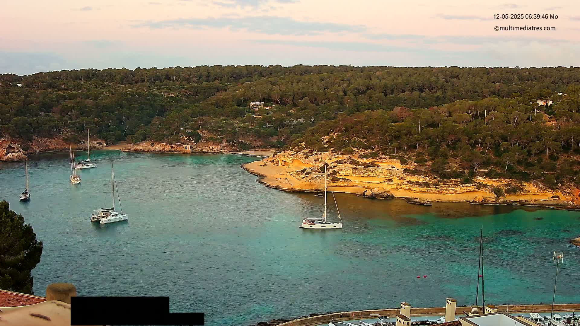 Several sailboats are anchored in a calm, turquoise bay surrounded by a lush, green, hilly coastline under a partly cloudy sky.