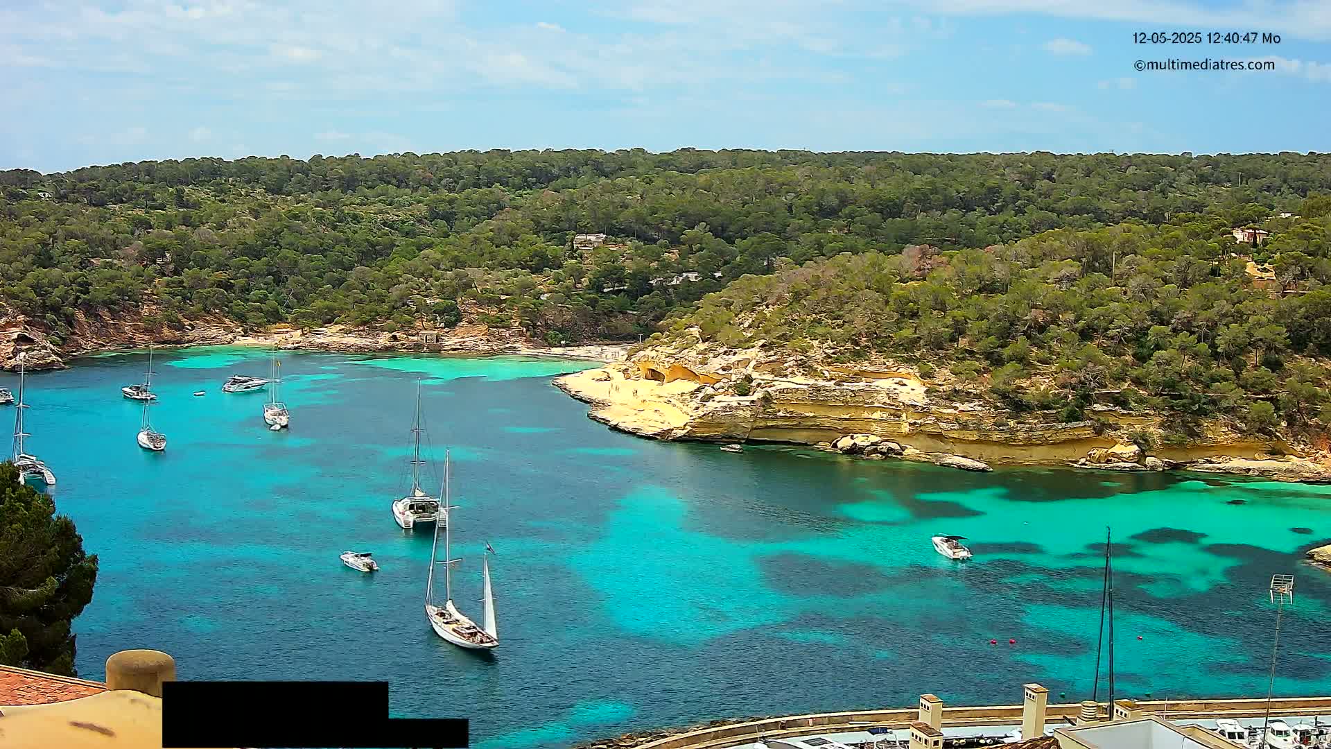 A picturesque cove with bright turquoise water is surrounded by lush green vegetation and sandy cliffs, dotted with several sailboats on a sunny day.