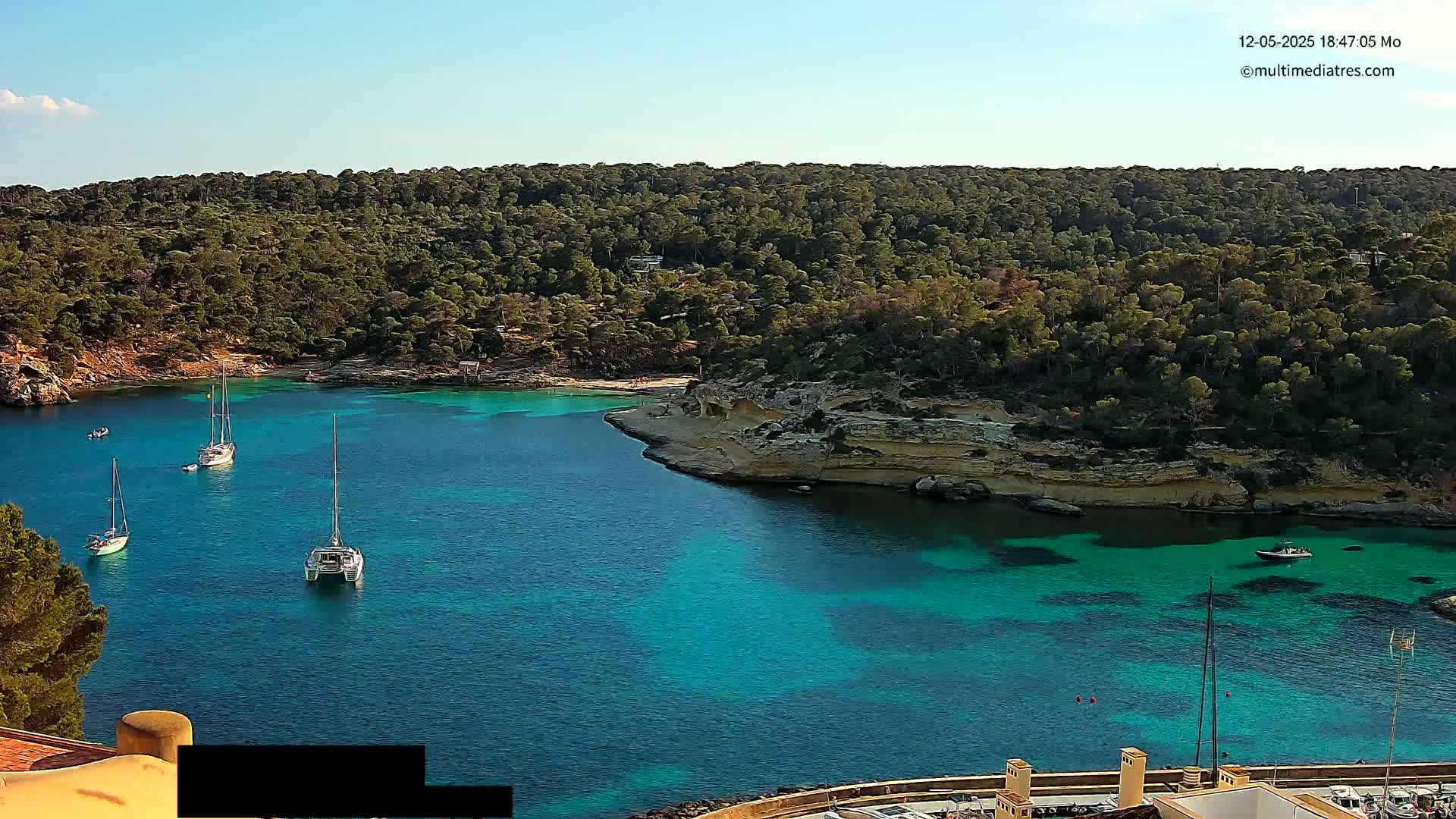A tranquil cove of bright turquoise water is nestled within a verdant, hilly landscape under a clear blue sky, with several sailboats anchored in the bay.