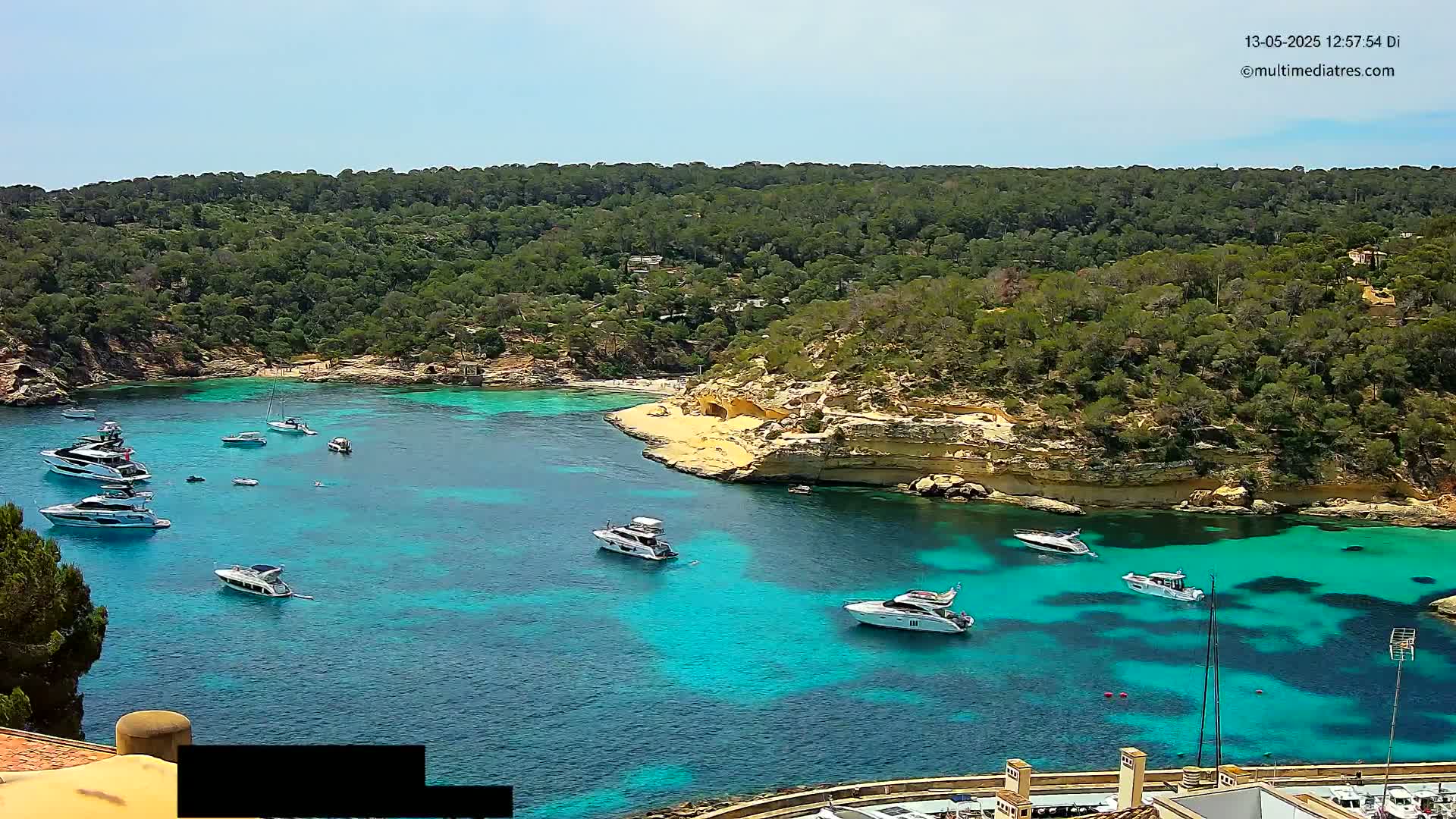 Several boats are anchored in a clear, turquoise bay surrounded by a lush green hillside under a bright, sunny sky.