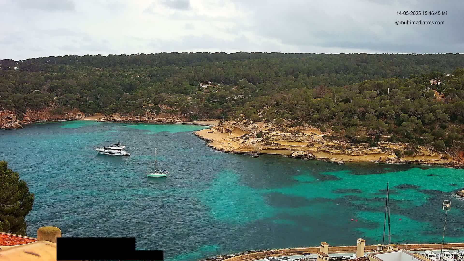 A cove with turquoise water is surrounded by a hilly, wooded landscape under an overcast sky; two motorboats and a sailboat are visible in the water.
