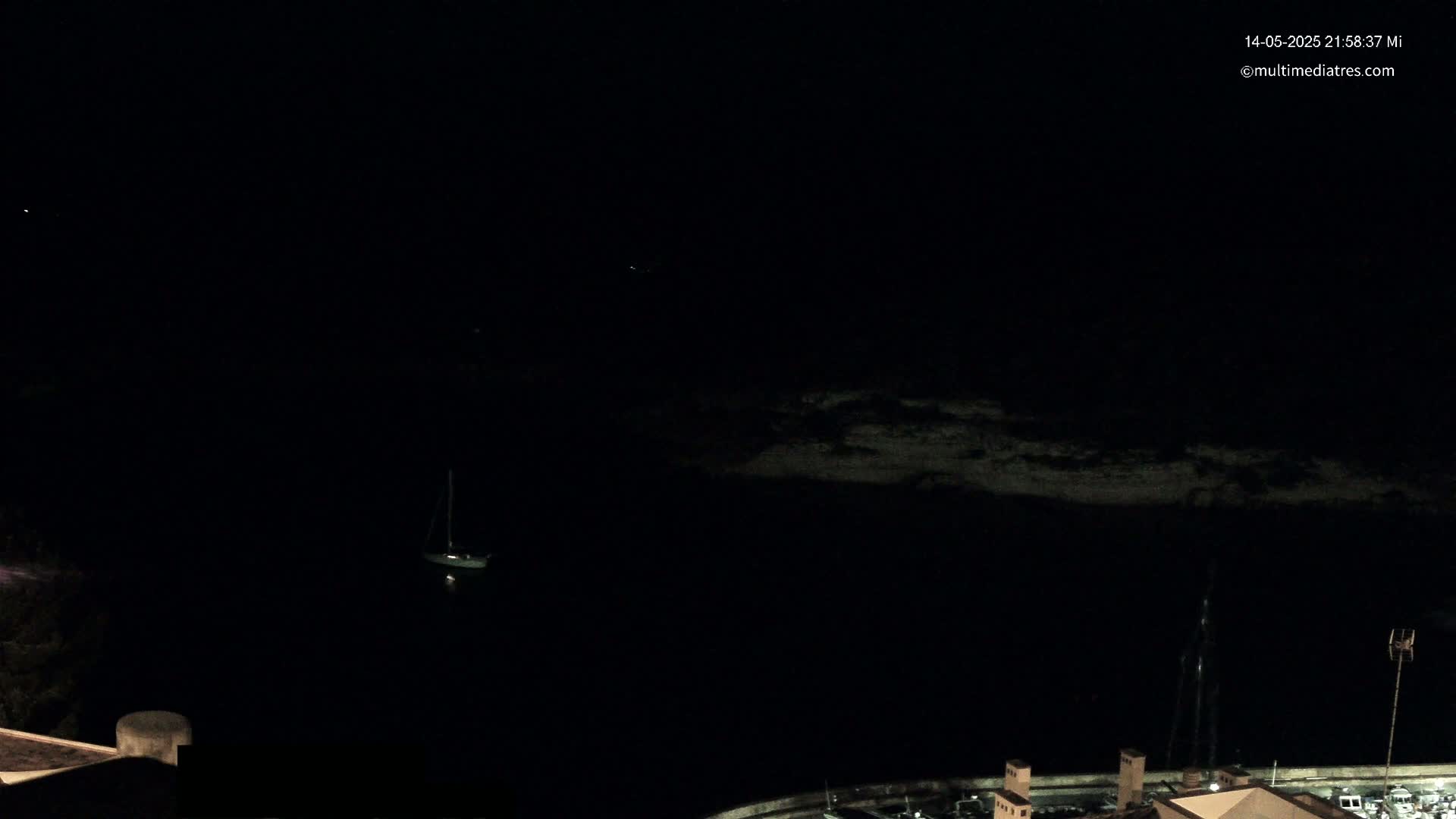 A dark, nighttime view of a sailboat at anchor in a calm body of water near a rocky shoreline and buildings.
