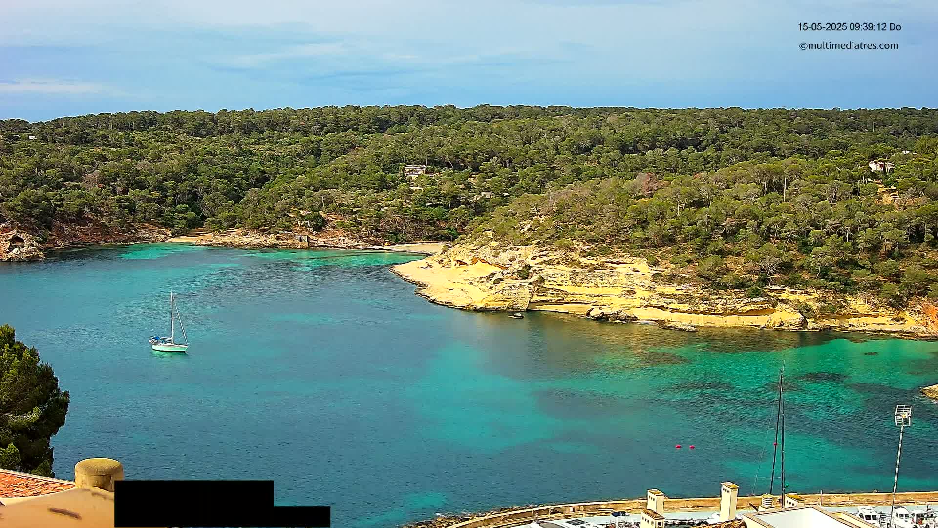 A sailboat is anchored in a calm, clear turquoise bay surrounded by a lush green forest and a pale yellow sandy beach under a mostly sunny sky.