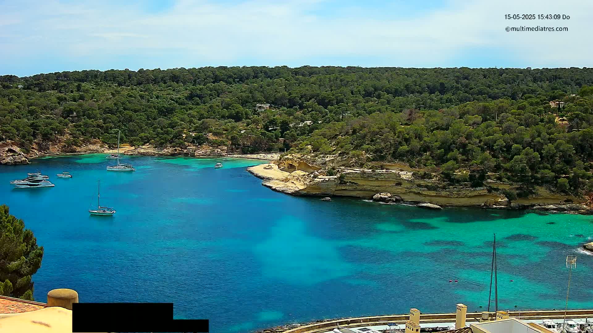 A tranquil cove with clear, turquoise water is surrounded by lush green vegetation and several boats are moored in the calm bay under a sunny sky.